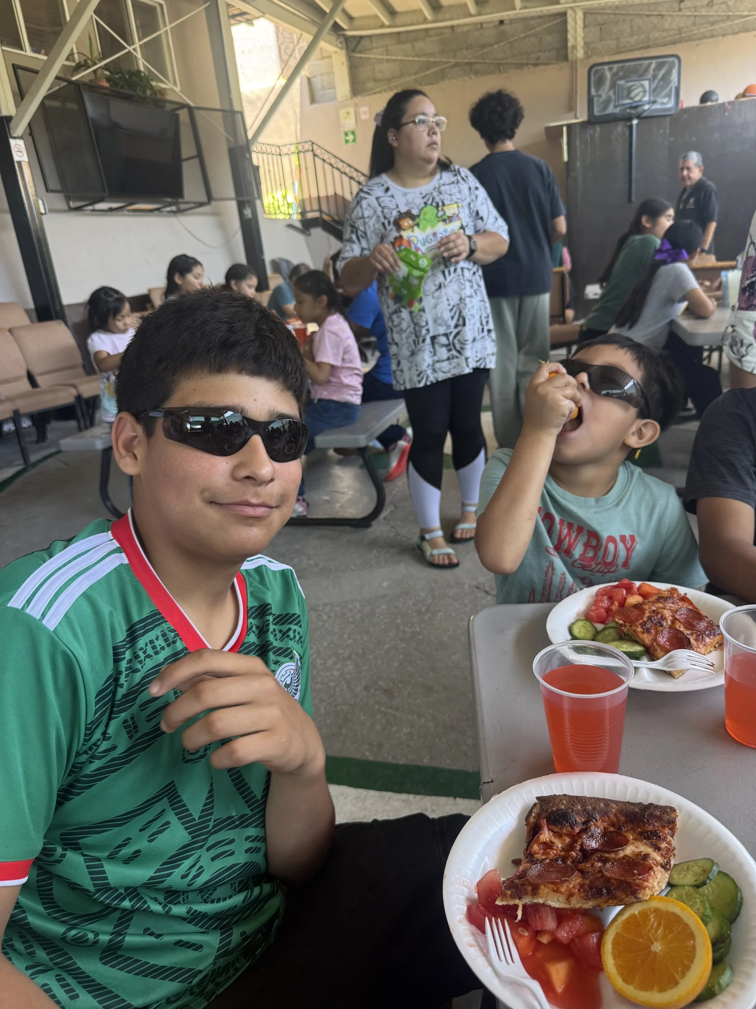 Young boy wearing sunglasses eating food at a table with pizza, fruit, and drinks in the foreground. In the background, a group of people including a woman holding a snack bag, seated and standing, inside a gym or recreational center.