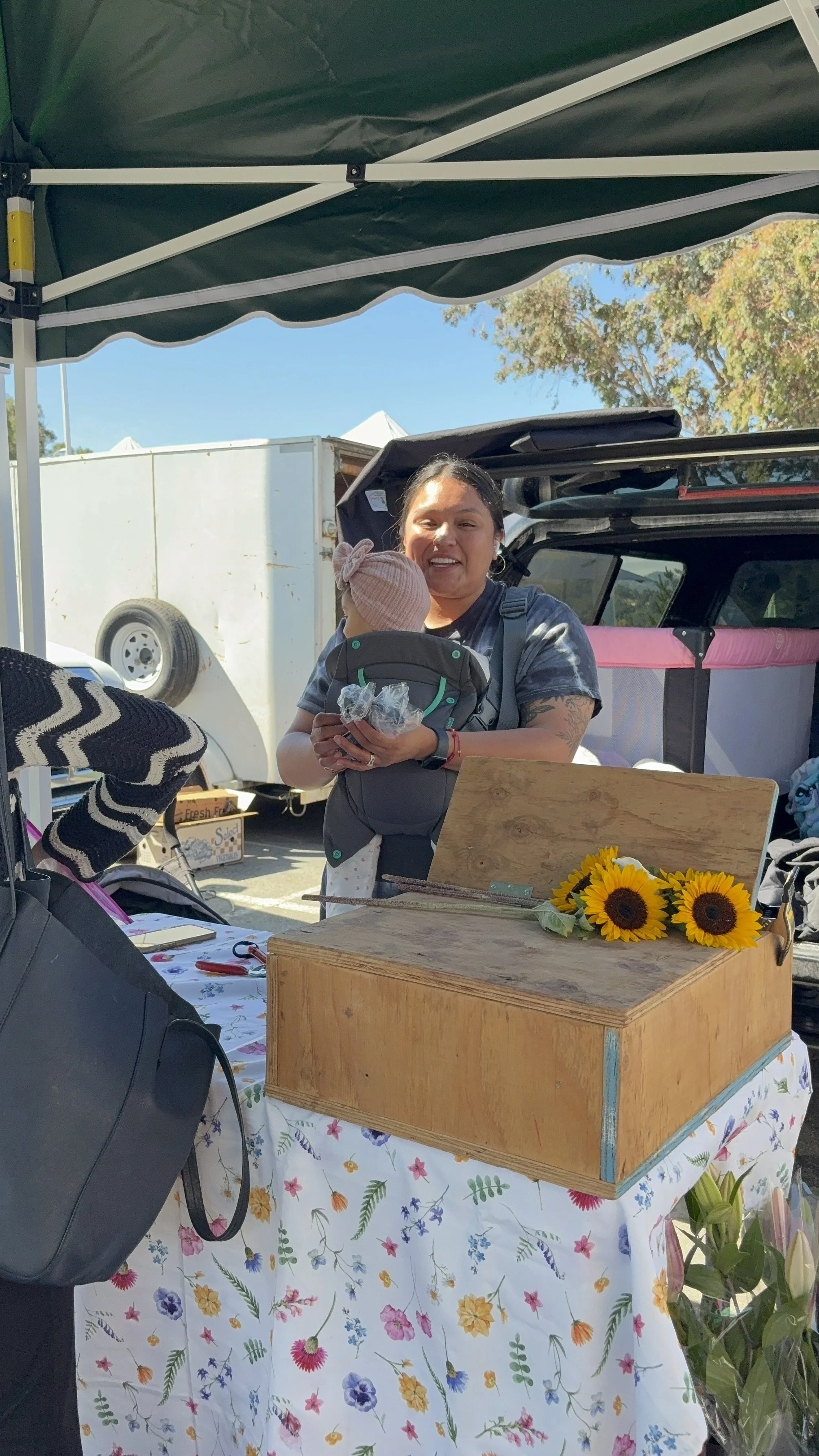 A woman standing behind a table at an outdoor market, holding a baby in a baby carrier, with sunflowers on a wooden box on the table, under a green canopy. There are trucks and trees in the background.