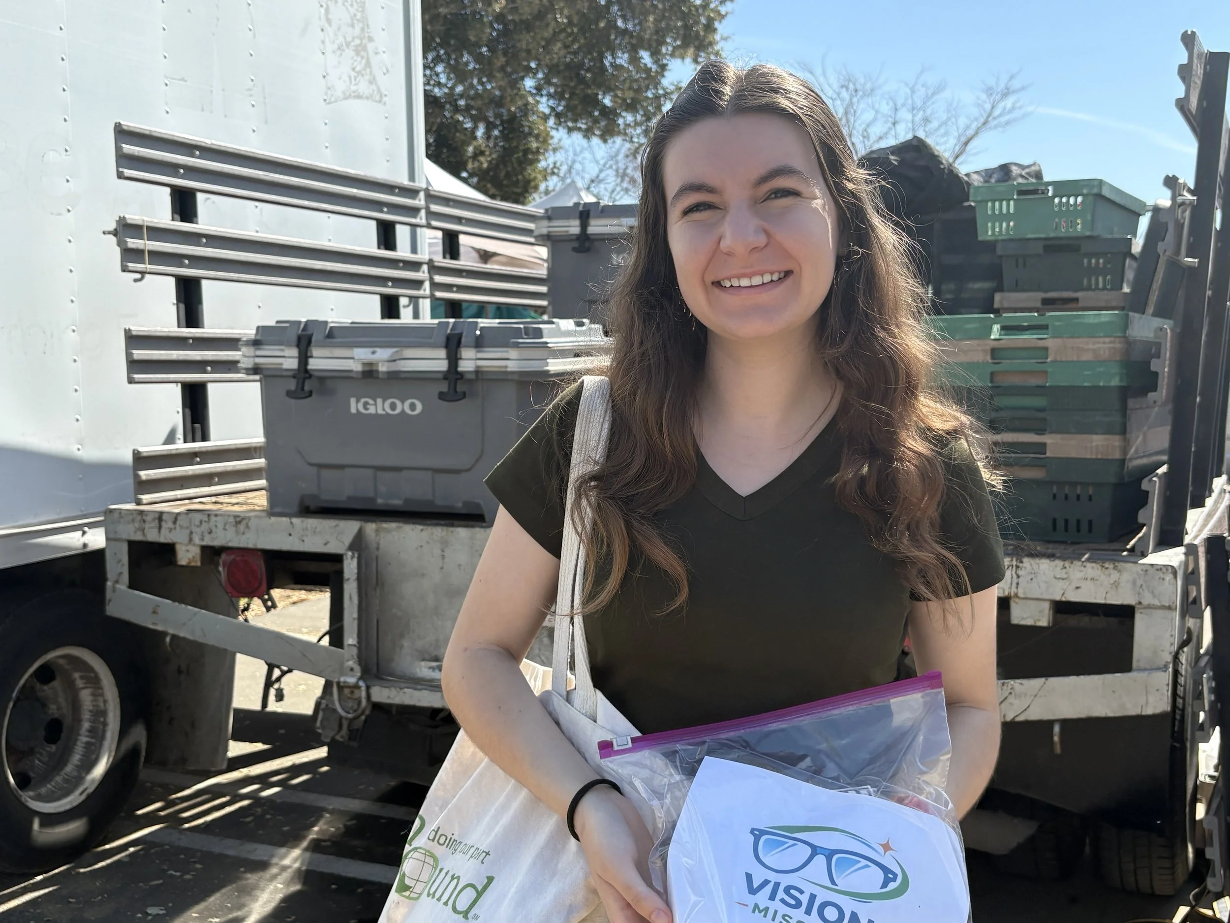 A smiling young woman with long brown hair standing outdoors in front of a truck loaded with plastic bins and metal shelving. She is holding a plastic bag and a paper bag with Vision Mission Logos on them.
