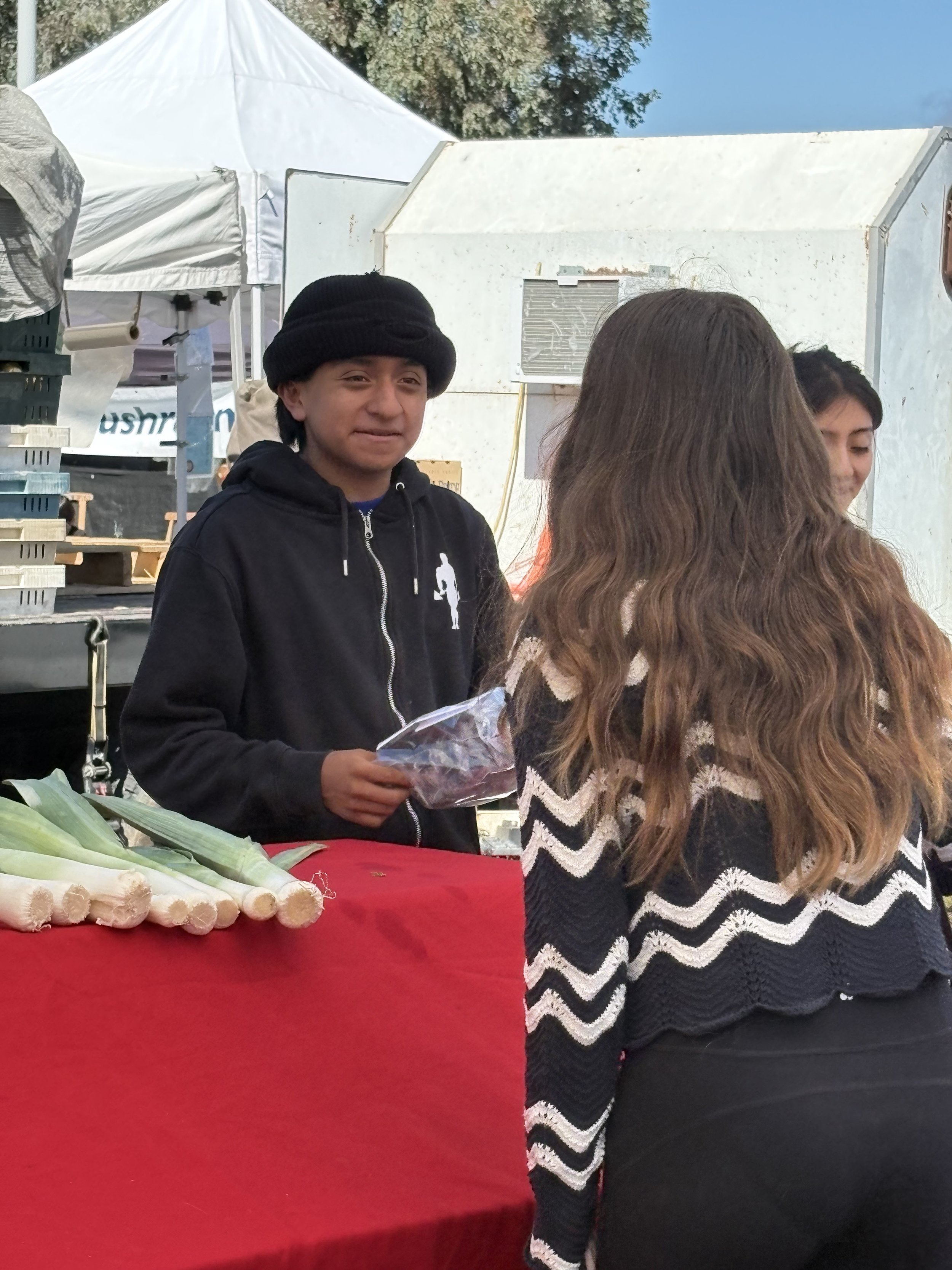 A young man in a black hoodie and black hat standing behind a red table with leeks and ginger at an outdoor market, talking to a woman with long, wavy brown hair wearing a black and white zigzag sweater.