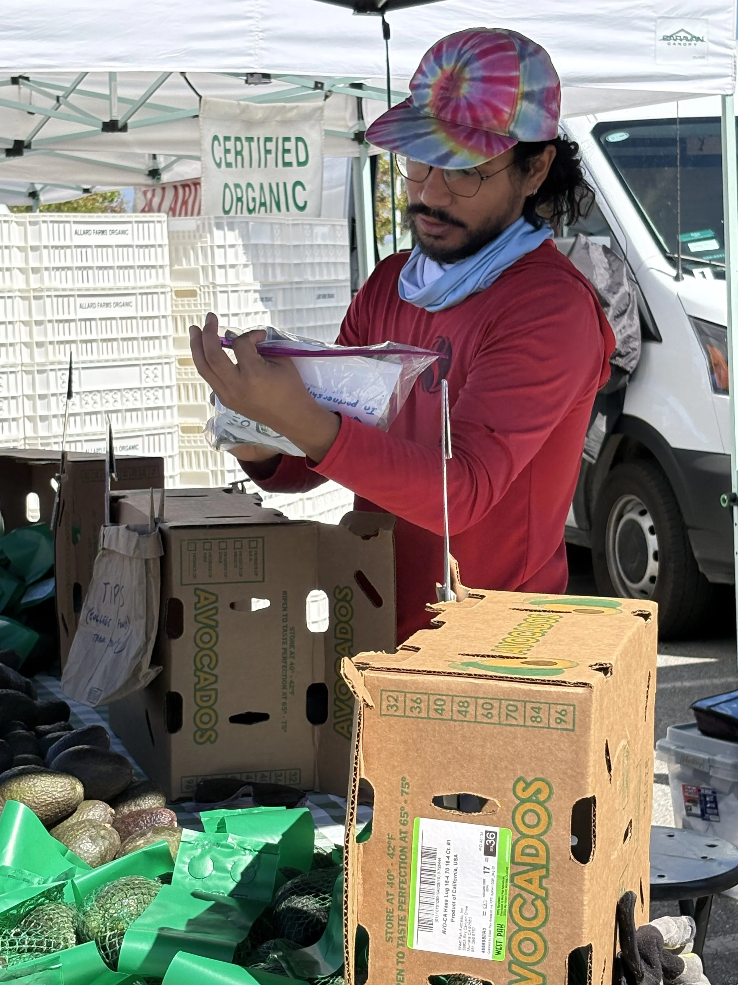 A man wearing a tie-dye hat, glasses, a red shirt, and a blue scarf around his neck shopping at an outdoor market. He is holding a plastic bag and looking at an item inside it. The market stall has a sign that says 'Certified Organic' and boxes of av
