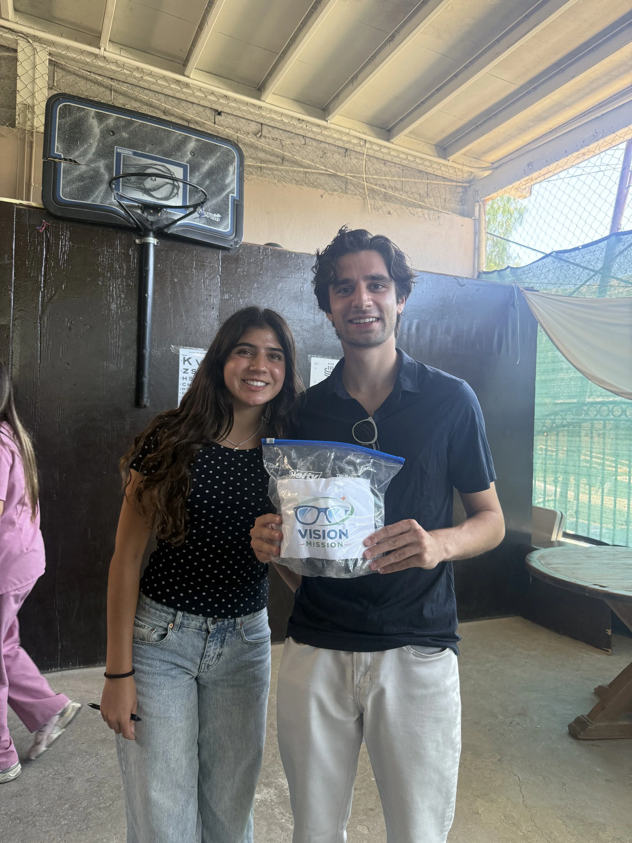 Group of two young adults smiling at a social event, with one holding a plastic bag with the logo 'VISION MISSION'. Background features a black wall, a small basketball hoop, and a netted outdoor area.