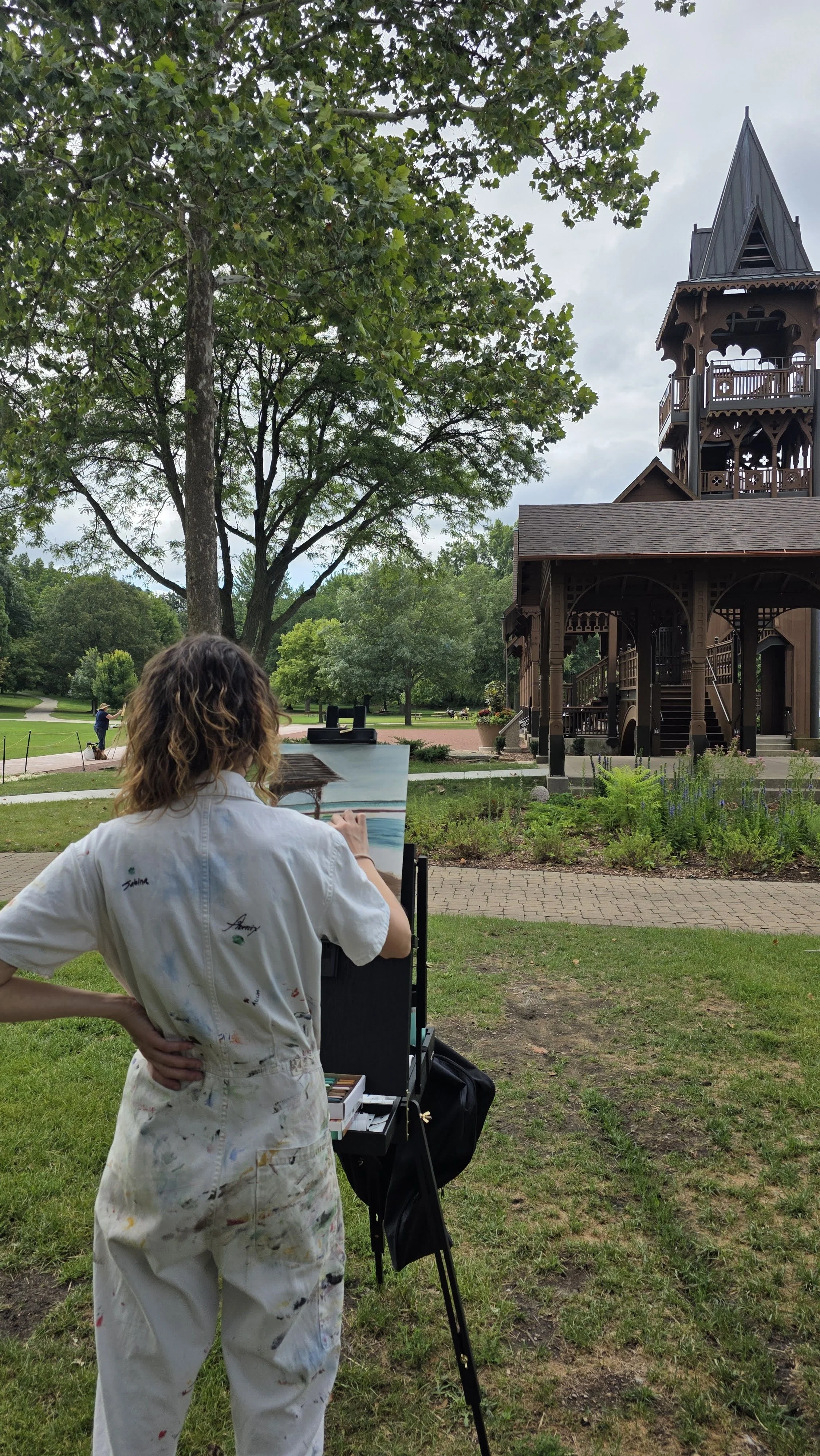 Person painting outdoors near a wooden building with a tower on a cloudy day.