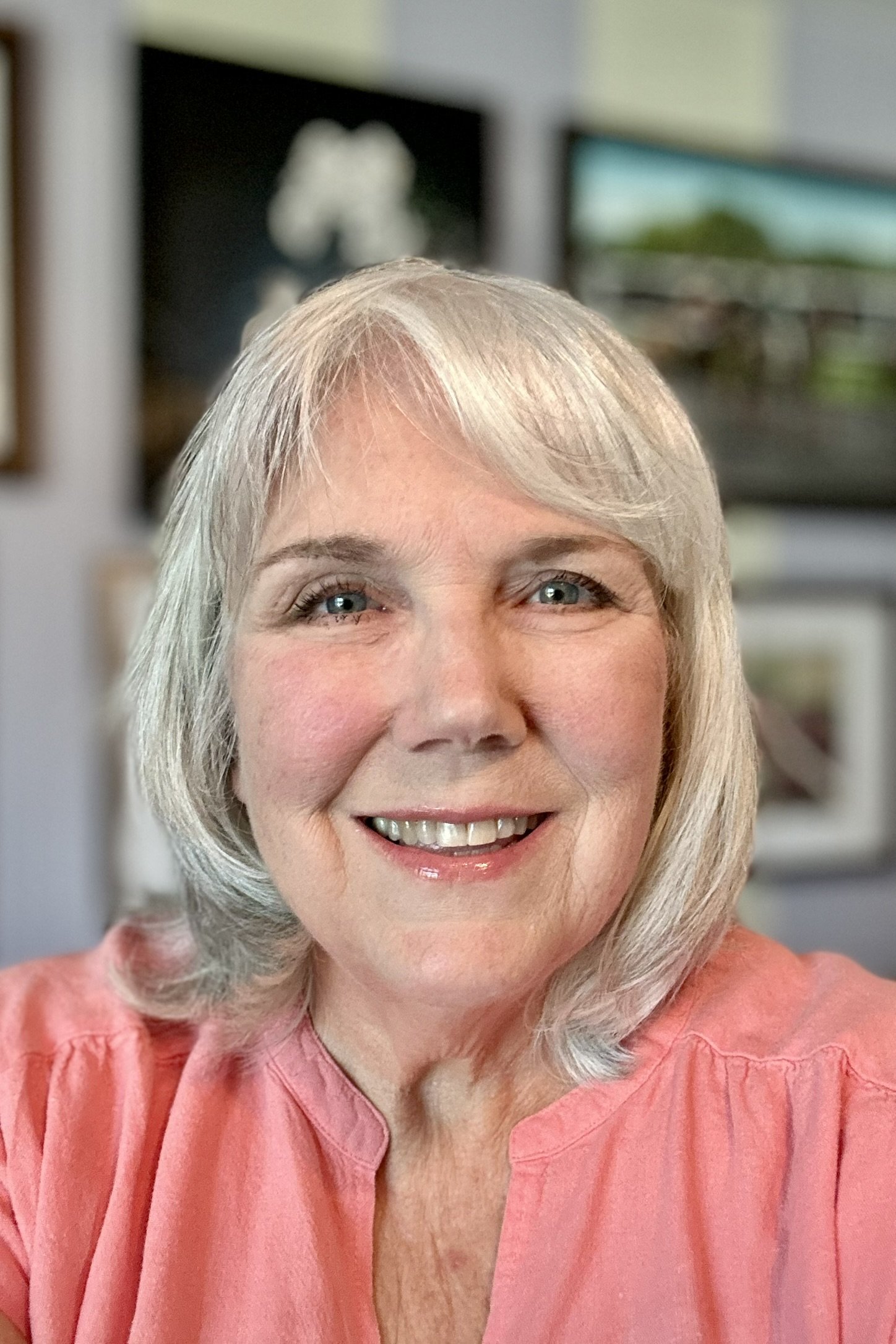 Close-up of a smiling older woman with silver hair, wearing a pink top, in a room with blurred background.