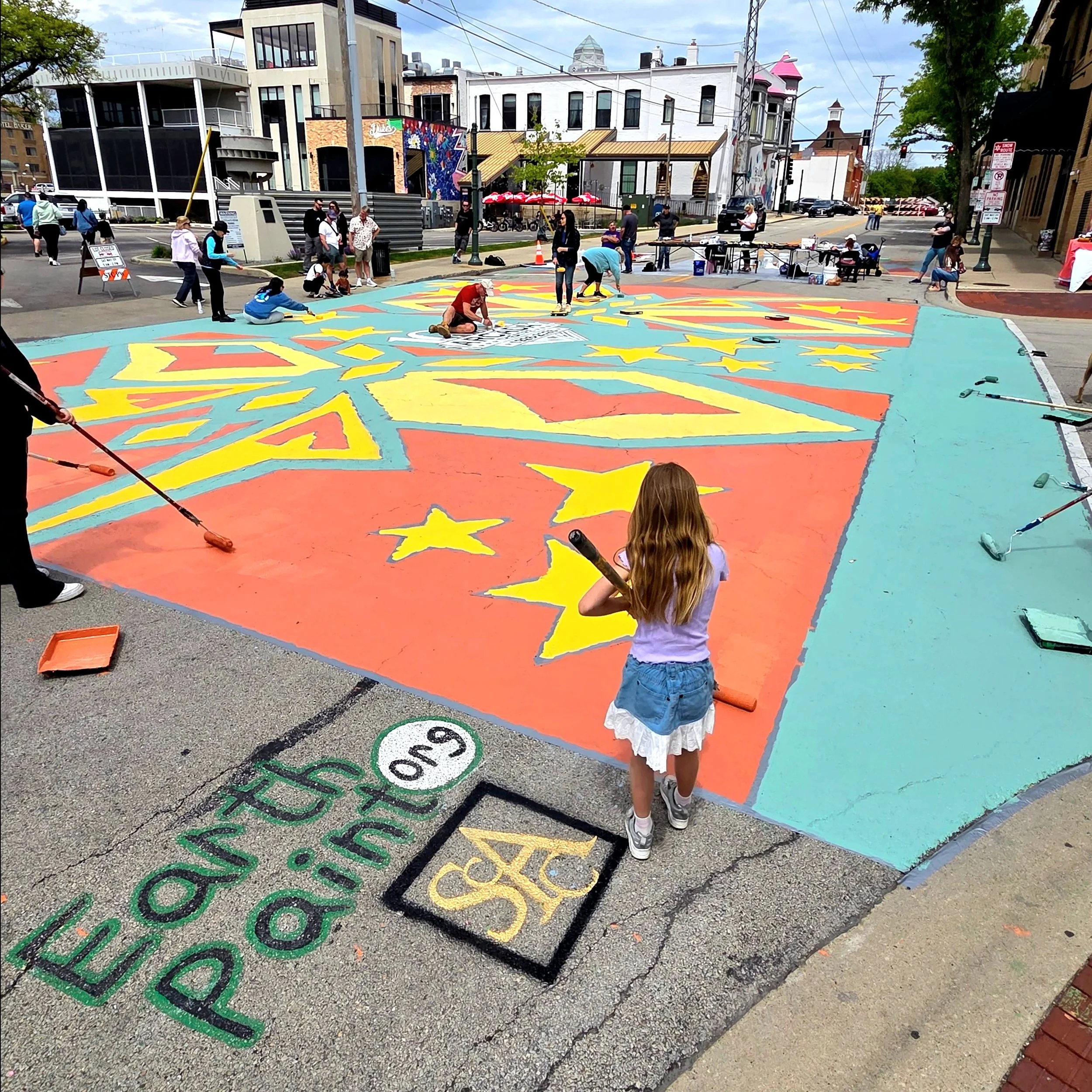 People creating a colorful mural on the street with various brushes and tools. Several onlookers are watching, some standing and others sitting. The scene is in an urban area with buildings and storefronts in the background.
