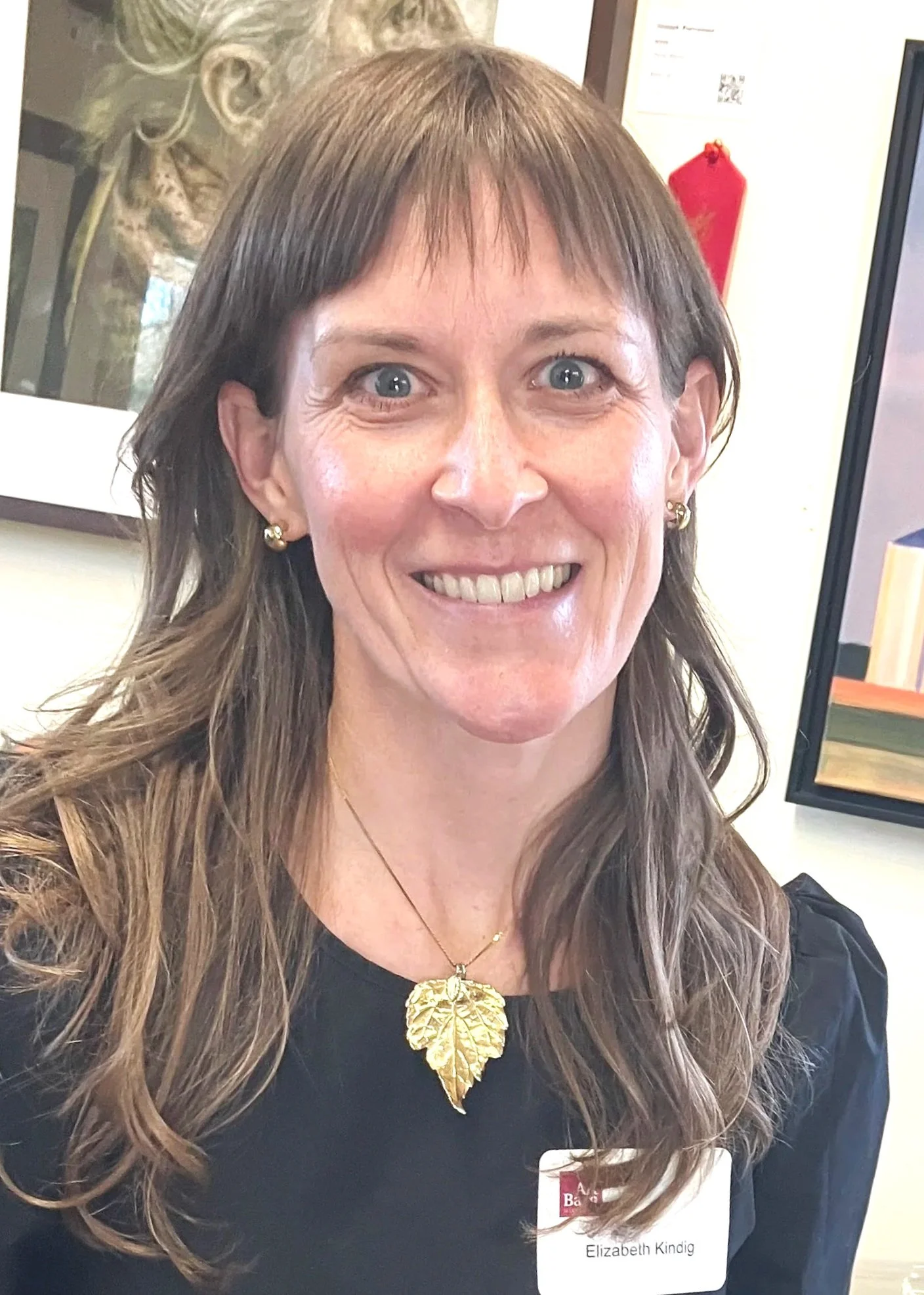 Close-up of a smiling woman with long wavy brown hair, wearing a black top, gold earrings, and a large gold leaf pendant necklace, with a name tag reading 'Elizabeth Kindig'.