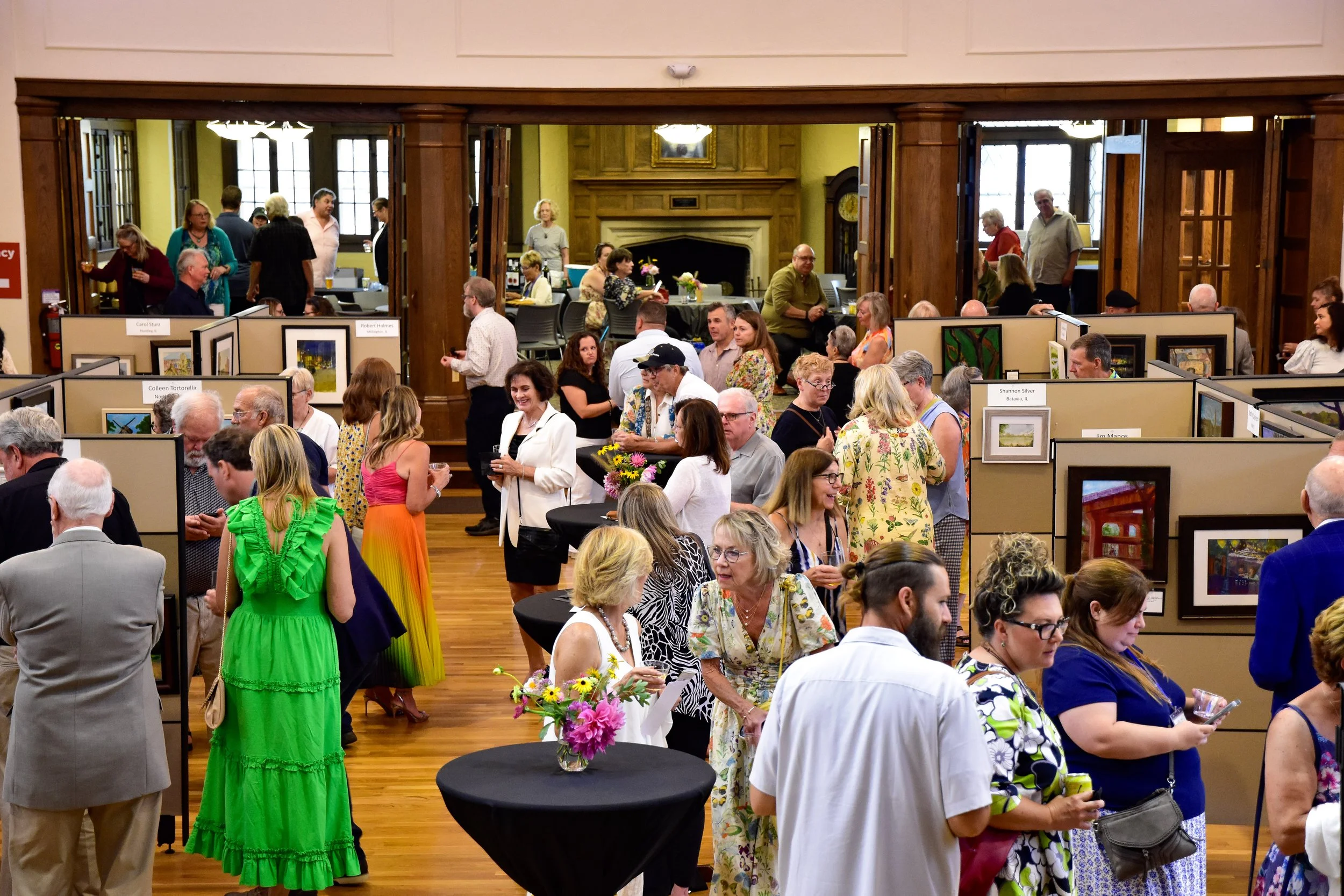 People attending an art exhibition in a gallery with paintings displayed on beige panels. The room has wooden floors, a fireplace in the background, and large windows letting in natural light. Visitors are conversing, looking at artwork, and some are holding drinks.