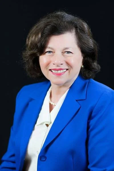 A woman with short brown hair wearing a blue blazer and a pearl necklace, smiling against a black background.