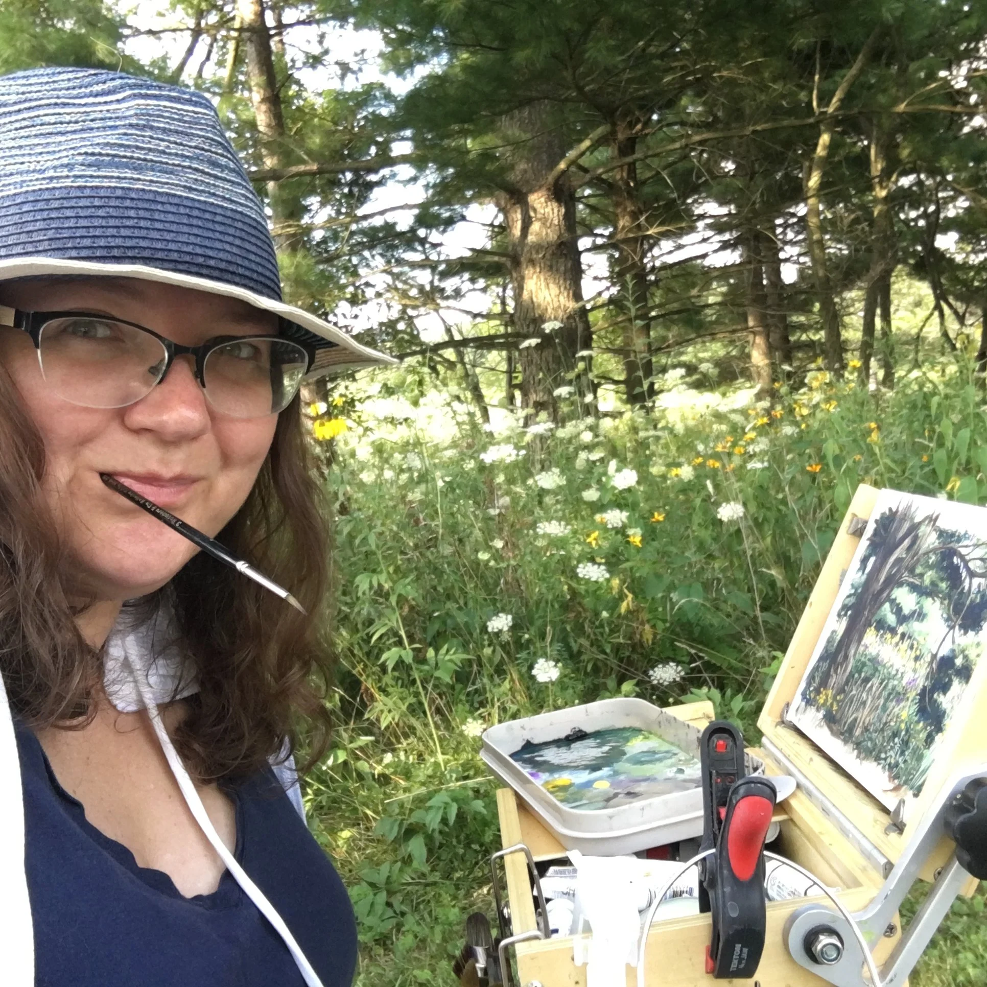 A woman wearing glasses, a striped wide-brimmed hat, and a dark shirt, appears to be working on a small woodworking project outdoors in a forest clearing. There are trees and yellow and white wildflowers in the background. She has a small table with tools, wood pieces, and a power tool.
