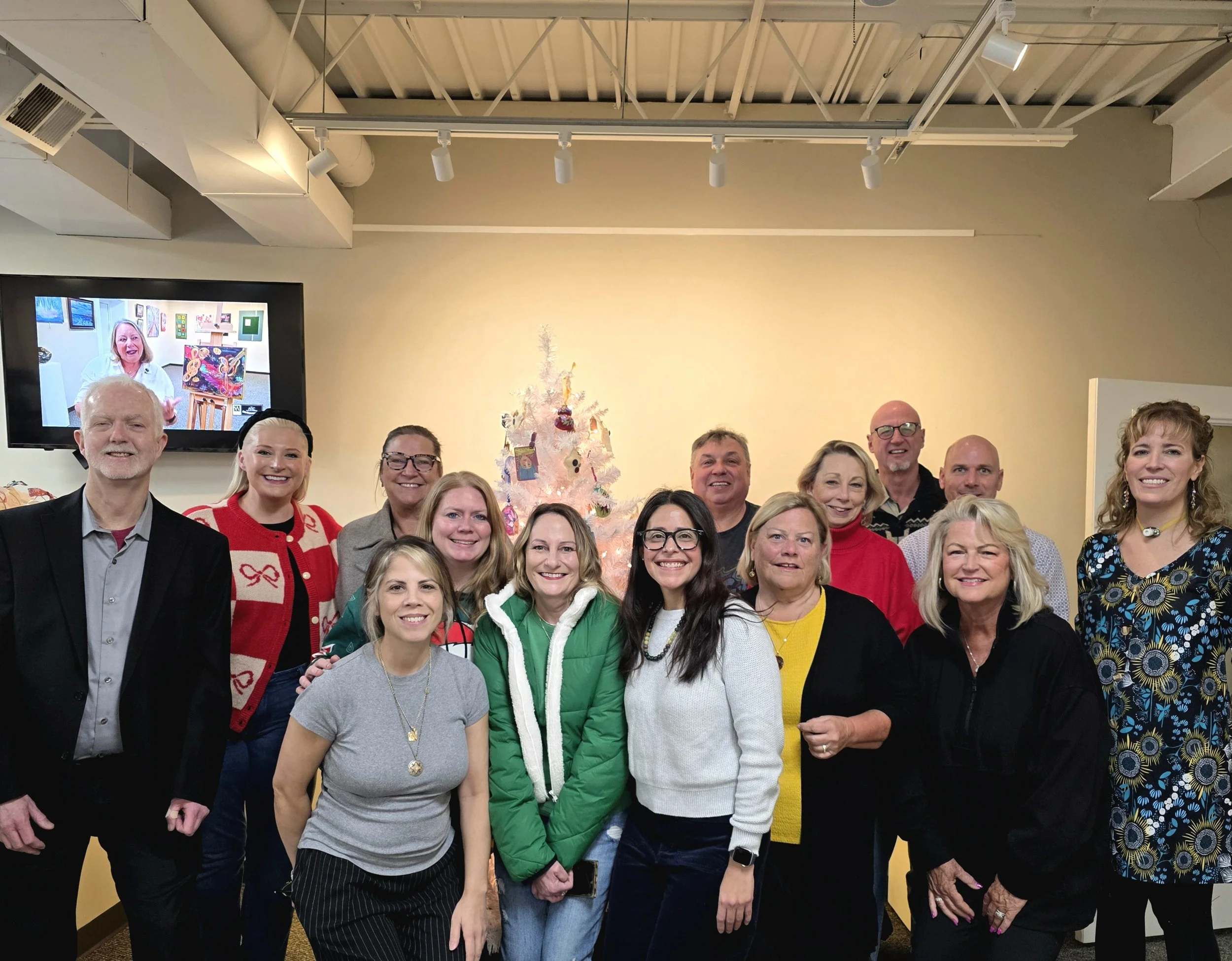 Group of fifteen people smiling in front of a Christmas tree with ornaments, during a festive gathering indoors. A large screen behind them displays a woman with colorful artwork.