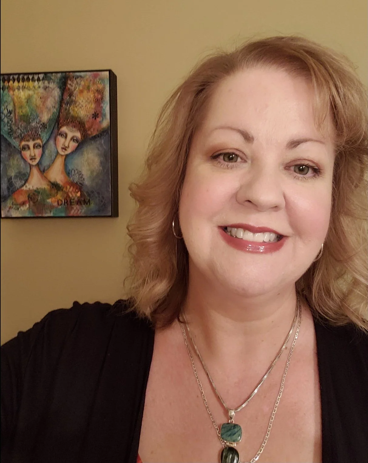 A smiling woman with earrings and layered necklaces in front of a colorful painting of two women with the word 'Dream' at the bottom.