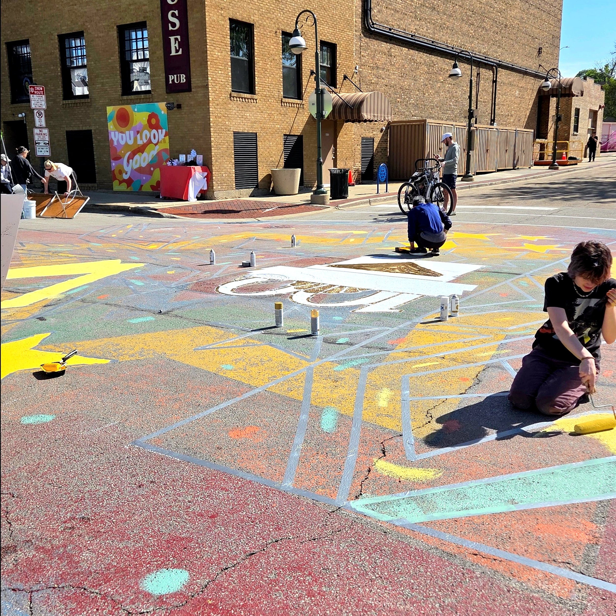 Colorful chalk art on the sidewalk outside a brick building with a sign that reads 'You Look Good.' Several people are working on the chalk art, some crouching and others standing, with spray paint cans and chalk visible.