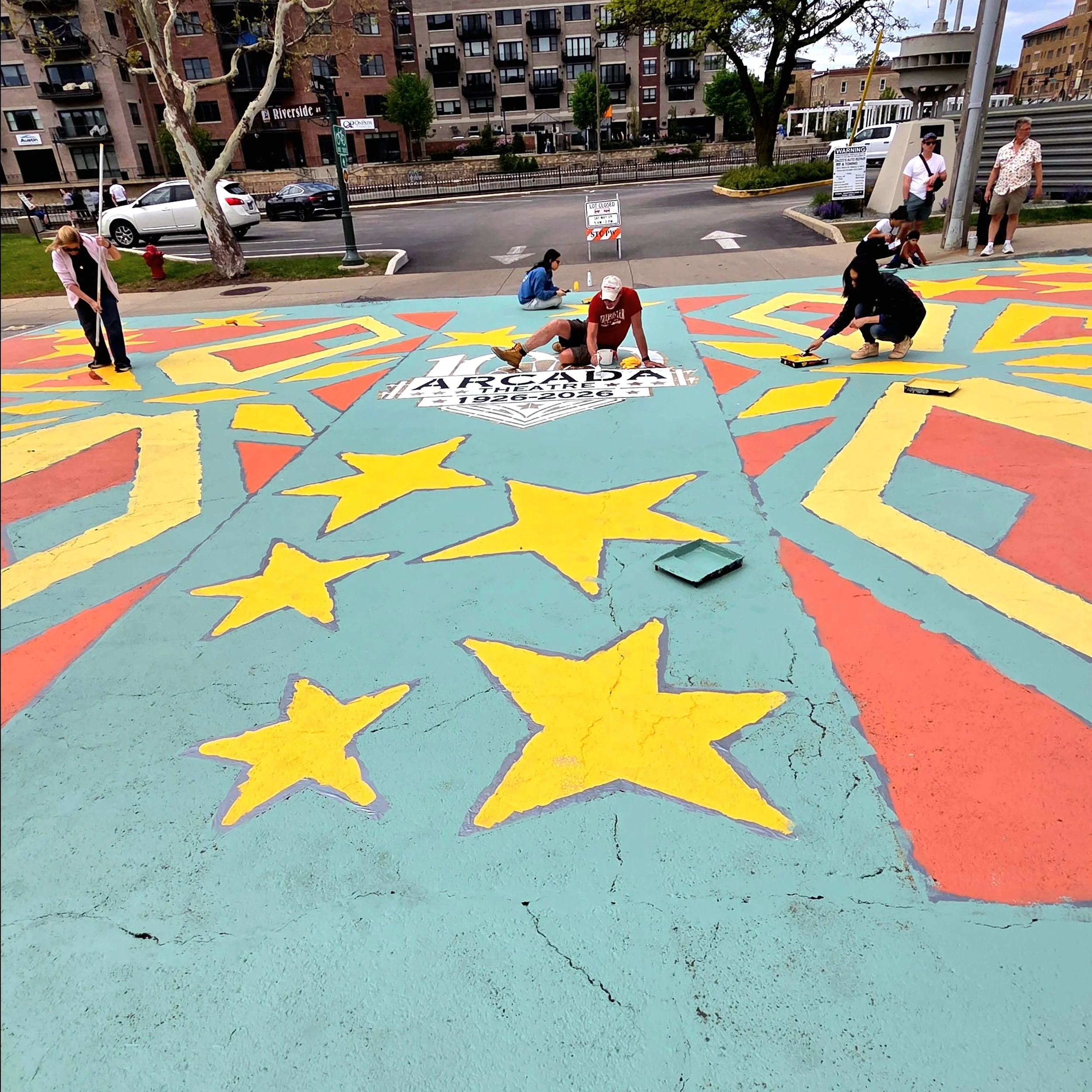 People creating a colorful mural on the pavement of a city sidewalk, with buildings and parked cars in the background.