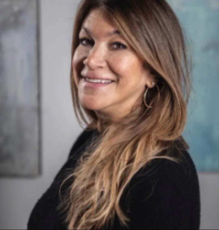 Woman with long, wavy brown hair, smiling and wearing hoop earrings, indoors with abstract artwork in the background.