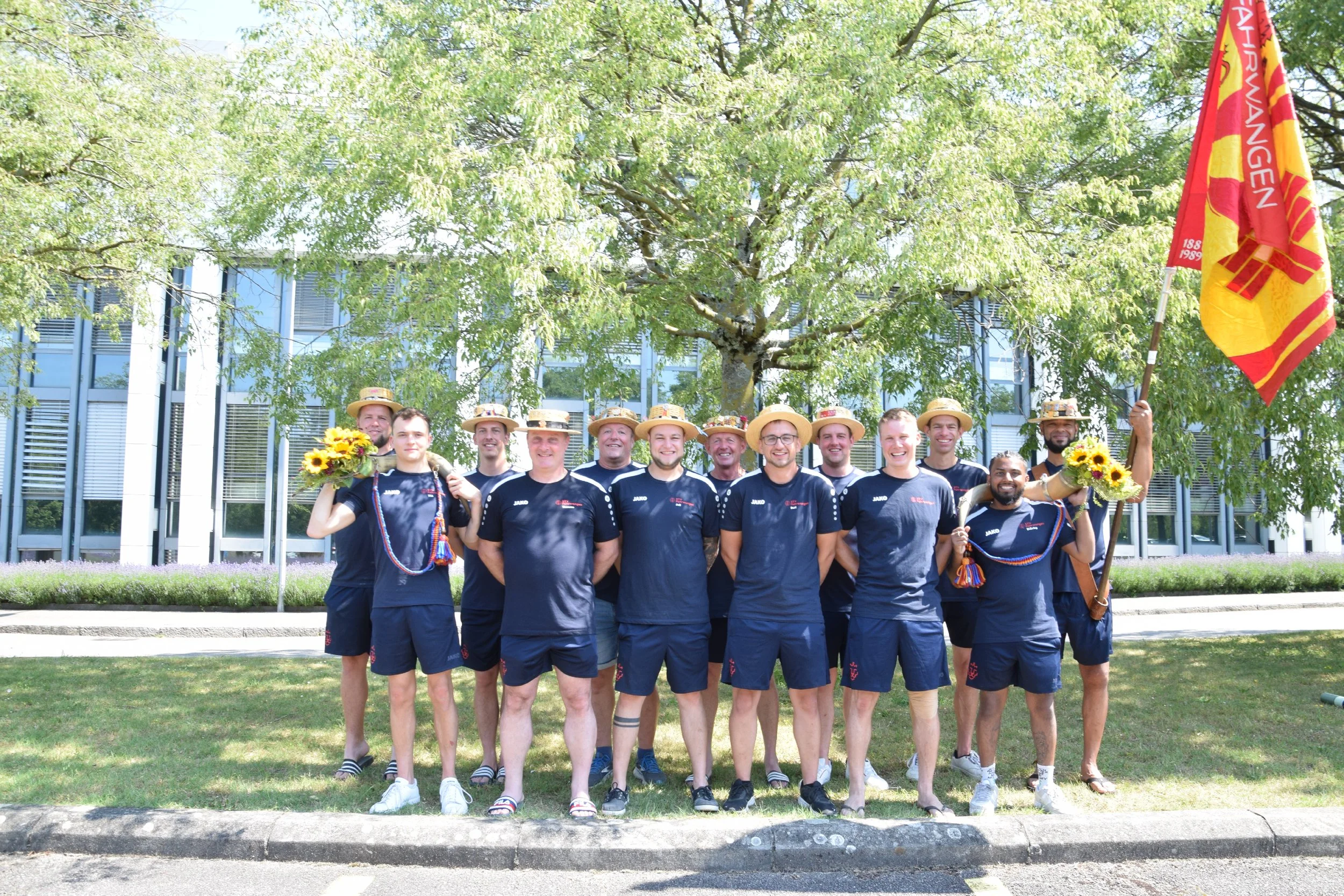 STV Fahrwangen in blauen Uniformen und Strohhüten, posieren vor einem Baum und einem modernen Gebäude, einer hält eine Flagge. Es ist sonnig, und die Turner freuen sich.