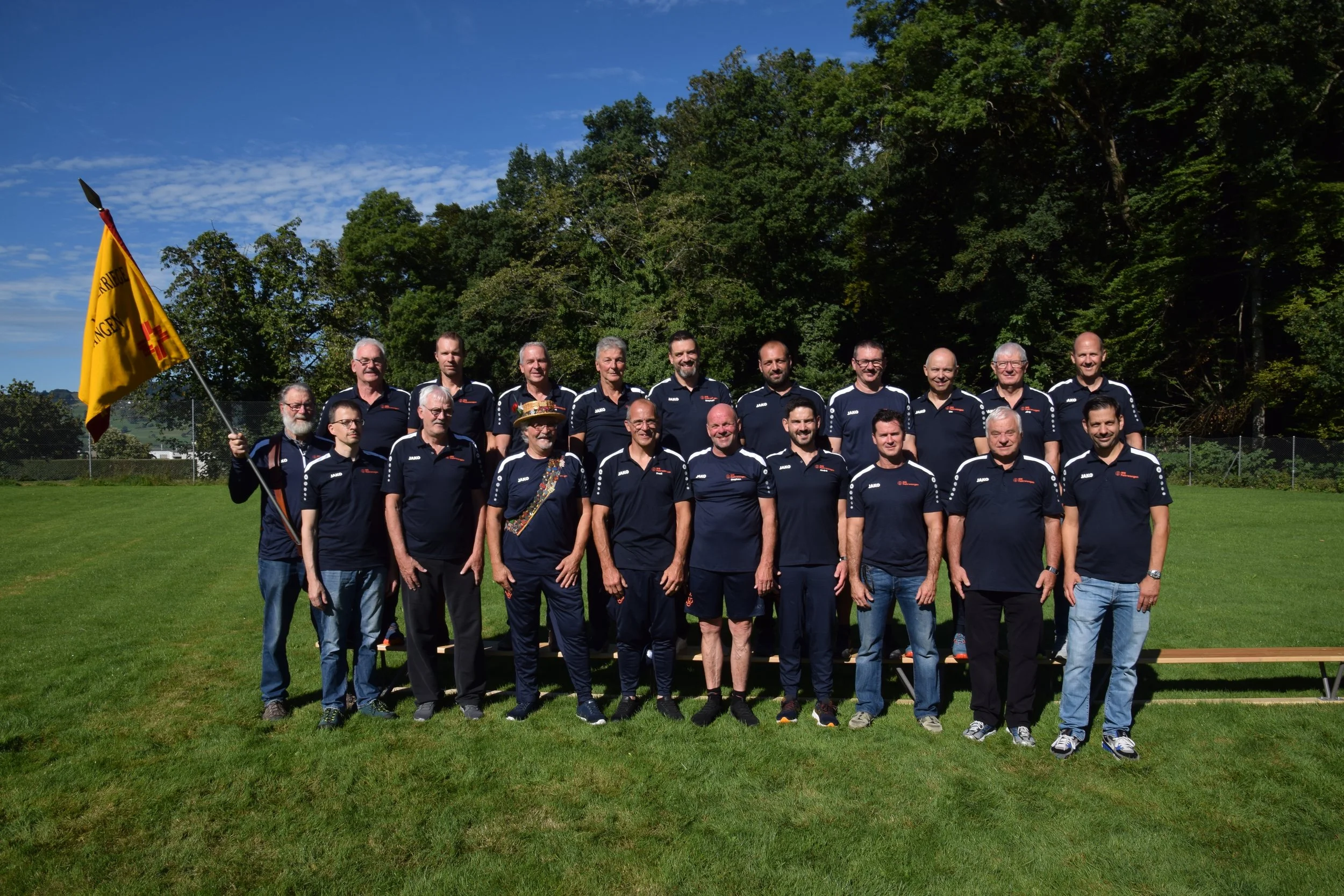 Männerriege Fahrwangen, MR, in schwarzen T-Shirts, stehend auf einem Rasen, mit einem Banner und einem Platz im Hintergrund, bei sonnigem Wetter.