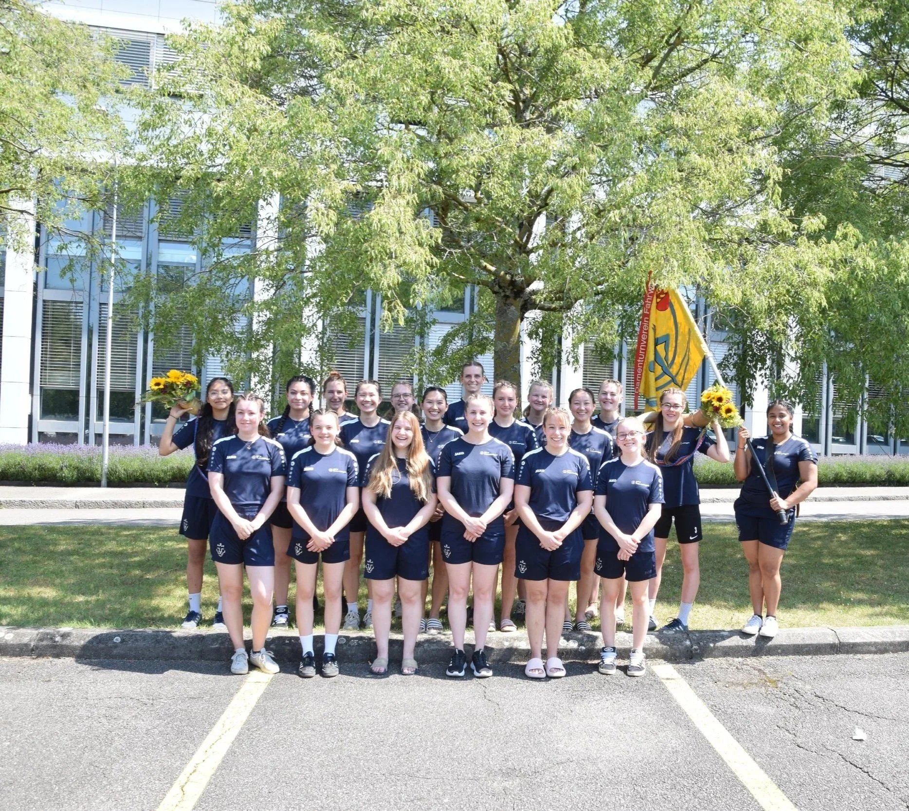 DTV Fahrwangen, in dunkelblauer Sportbekleidung steht vor einem Baum im Freien, einige halten Sonnenblumen und eine trägt eine Flagge. Sie posieren für ein Gruppenfoto.