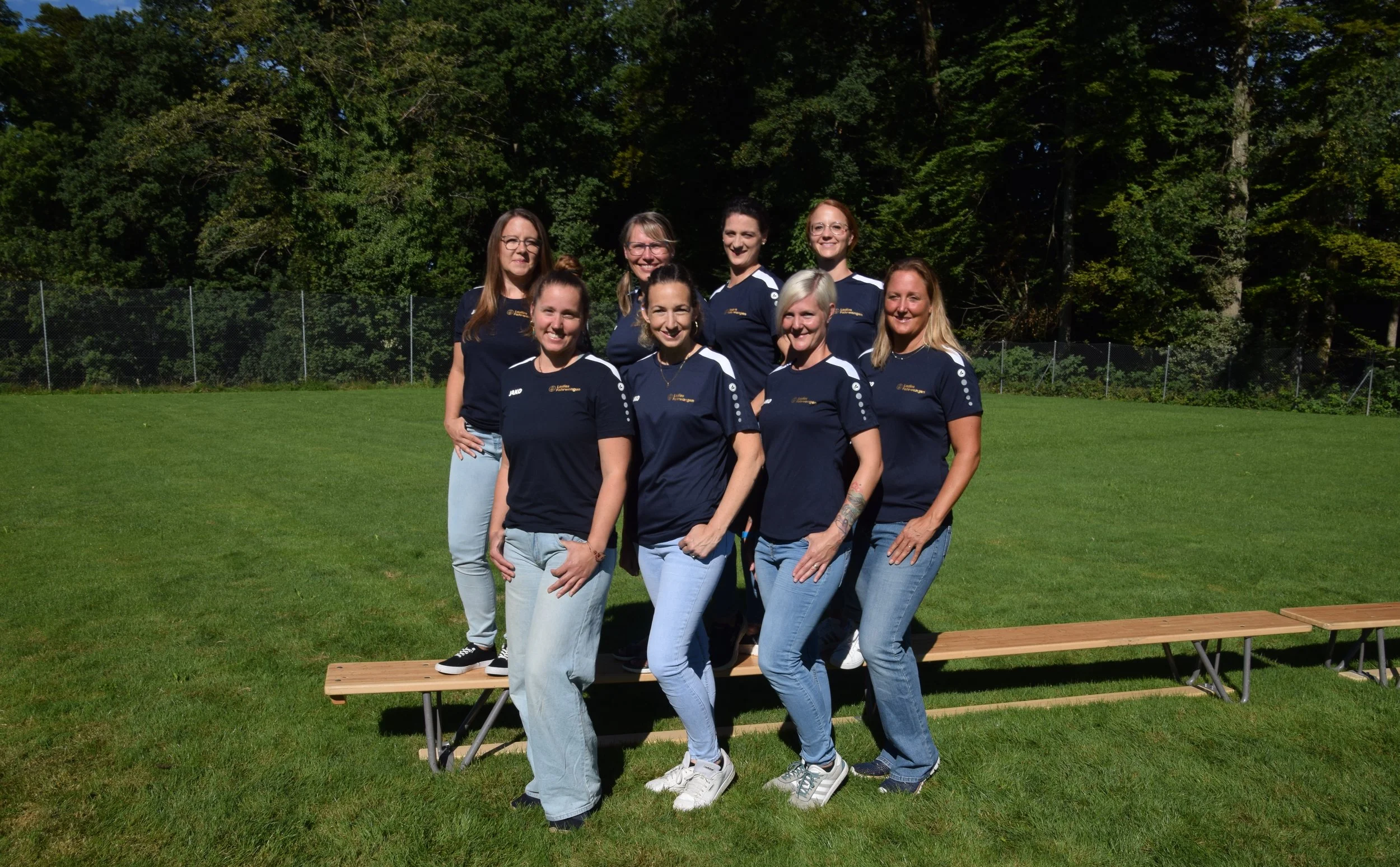 Ladies aus Fahrwangen, die auf einer Holztribüne im Freien stehen, alle tragen dunkelblaue T-Shirts und Jeans, vor einem grünen Baumhintergrund bei sonnigem Wetter.