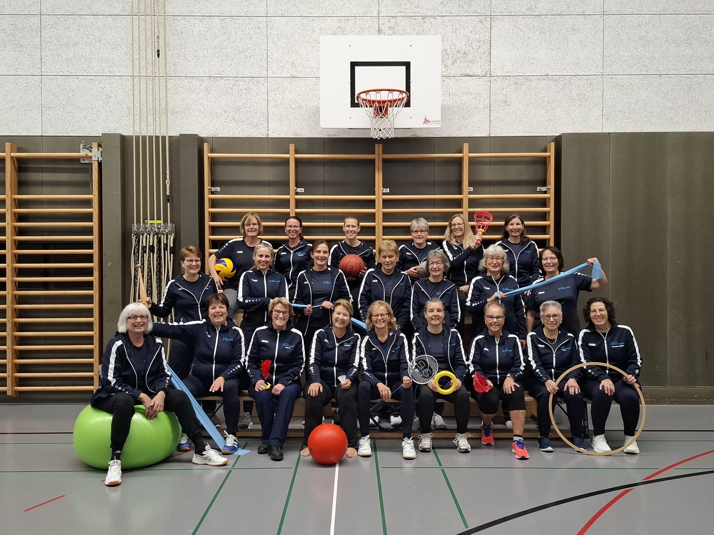FTV Fahrwangen in Sportkleidung, posieren in einer Turnhalle mit verschiedenen Sportgeräten, darunter Bälle, Hula-Hoop-Reifen und Seile.