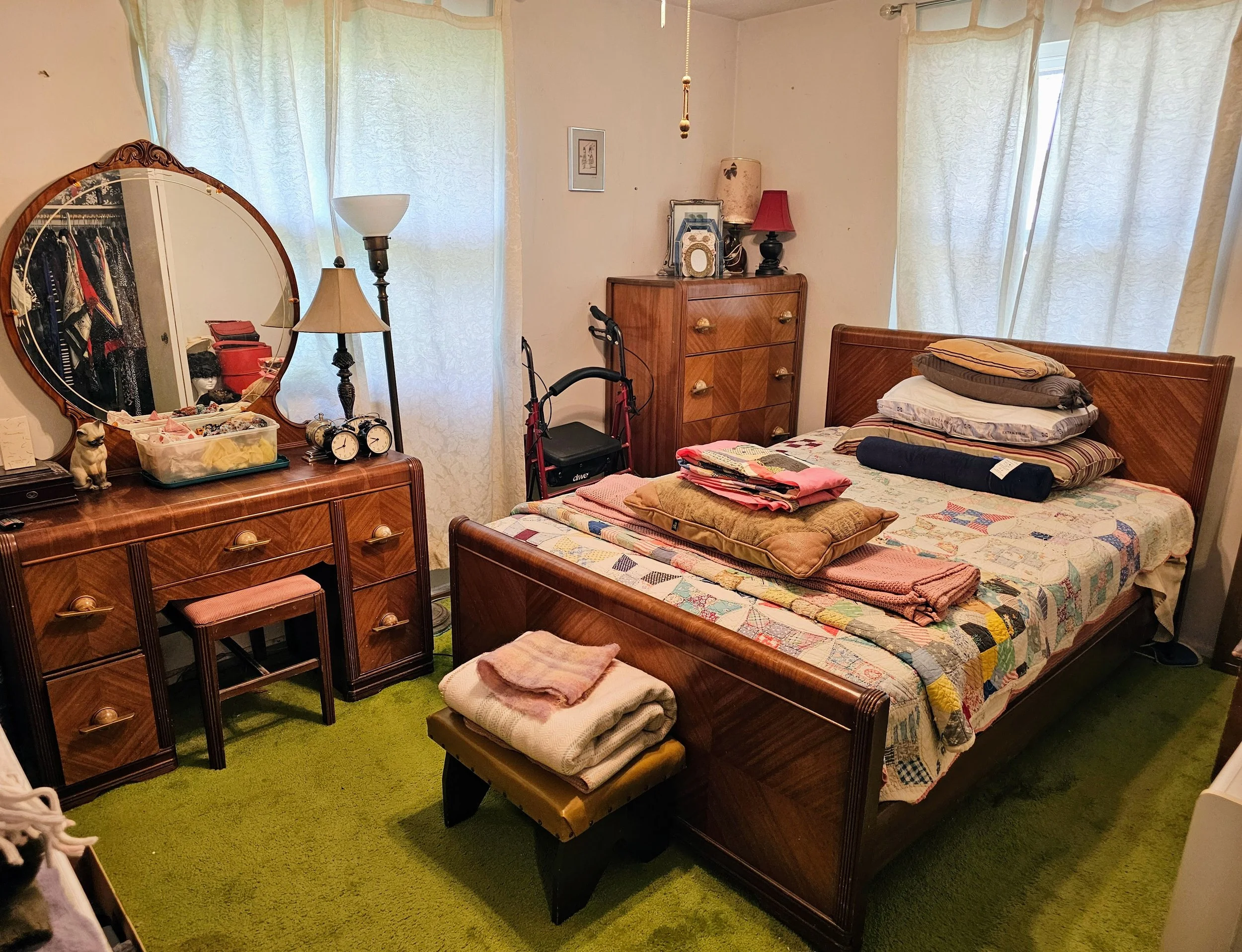 A bedroom with a wooden bed and a patchwork quilt, with pillows and folded clothes on top. A matching wooden dresser with a mirror and various items, a small bench with folded towels, and a bedroom vanity with a mirror, clock, and small decorative dog figurine. There's a window with light curtains and a green carpet covering the floor.