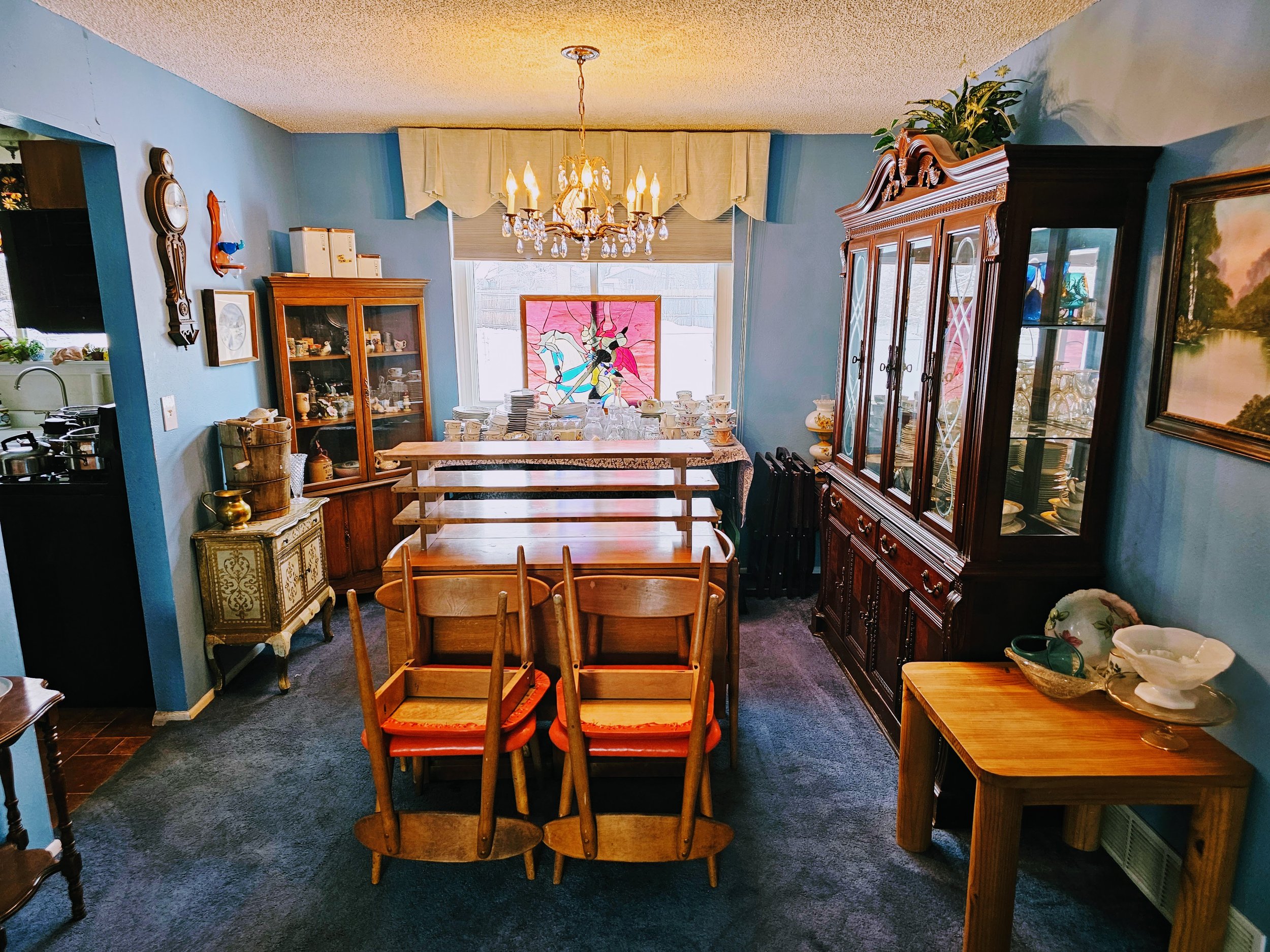 A dining room with blue walls, wooden furniture, and a chandelier. The table is set with dishes and a stained glass window decorates the background. There are china cabinets and a side table with decorative items.