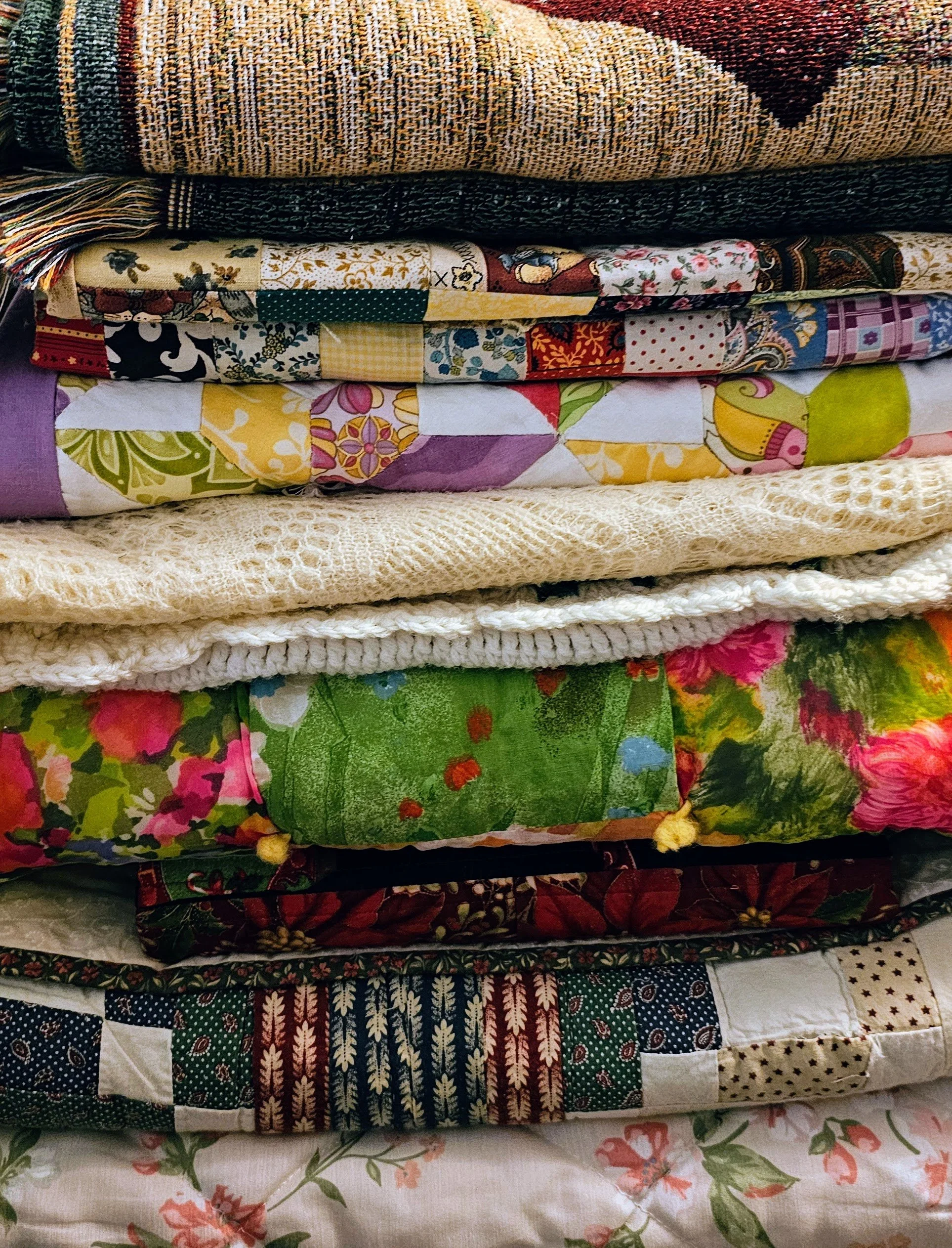 Stacked multicolored quilts with various patterns, including floral, geometric, and patchwork designs.