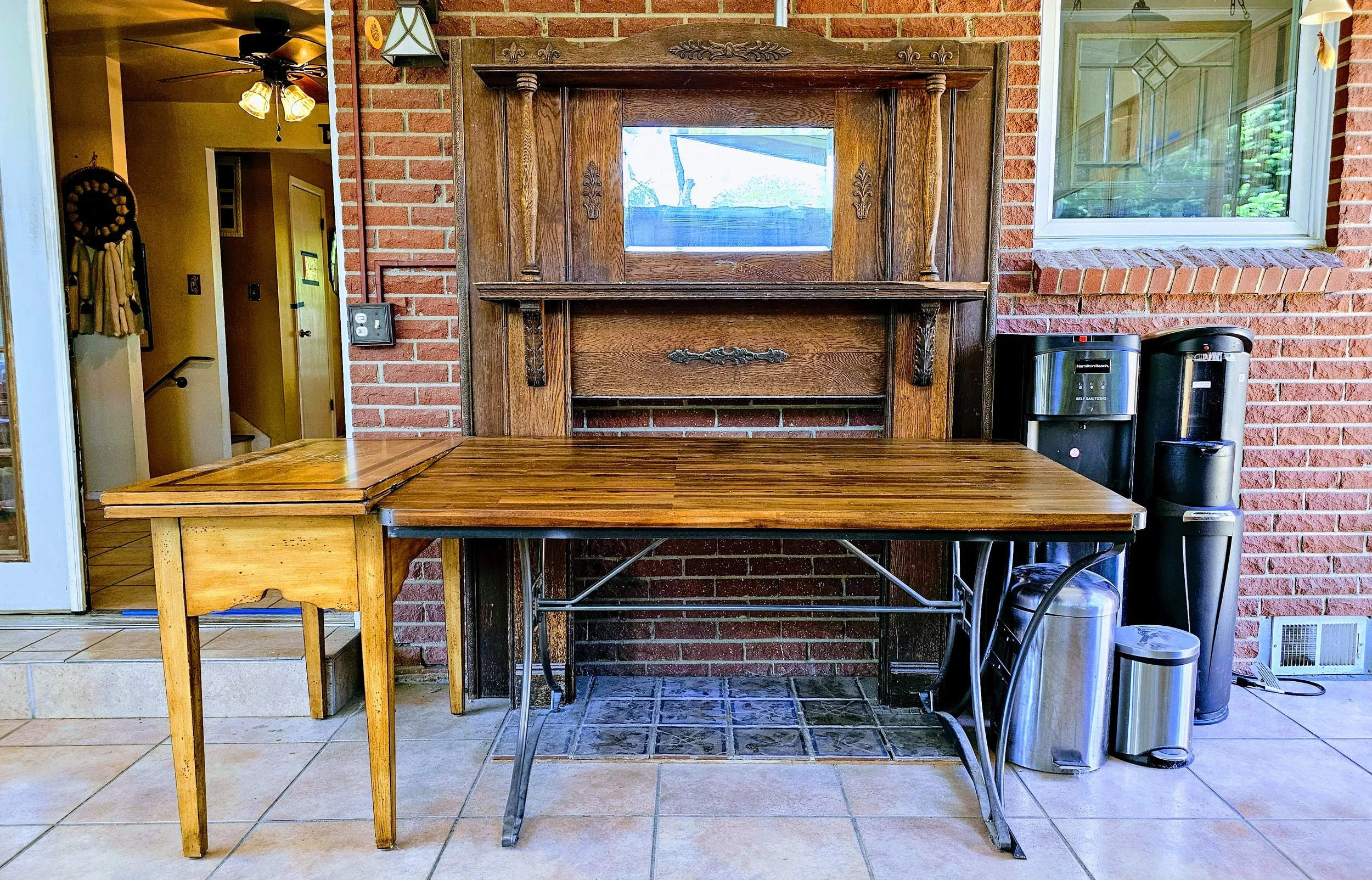 Outdoor patio area with a wooden table, vintage wooden cabinet with mirror, brick wall, water dispensers, and tiled floor.
