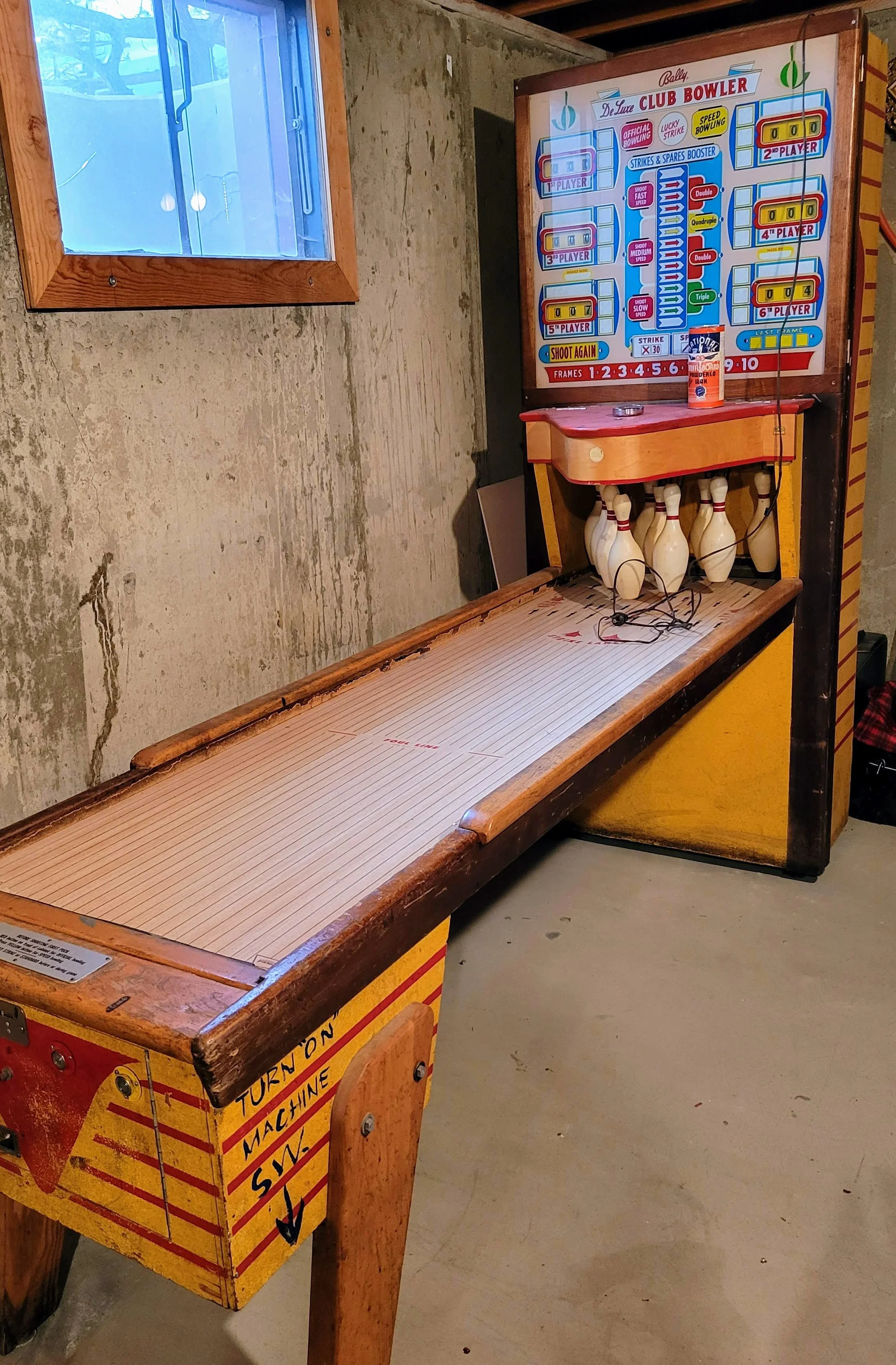 A vintage pinball machine with a wood finish, labeled as 'De Luxe Club Bowler', featuring a bowling-themed design and mechanical parts for gameplay, set against a concrete wall with a small window.