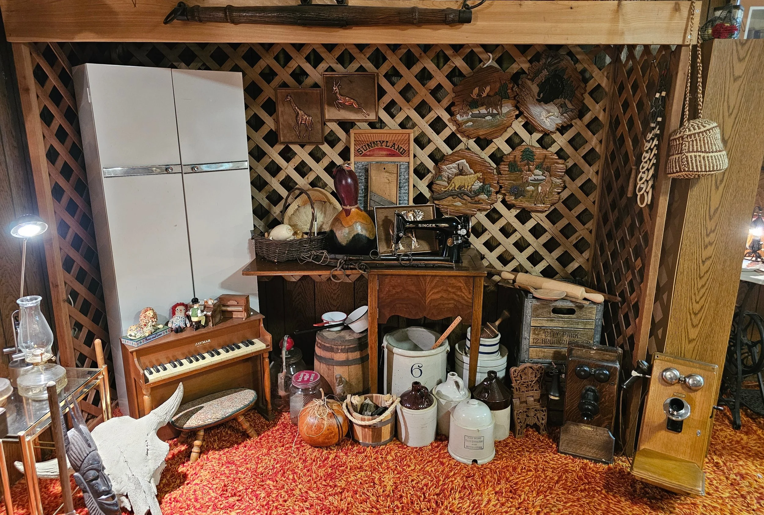 Vintage decor with a wooden lattice wall, a white refrigerator, small decorative figurines on a miniature piano, a sewing machine, various jars and containers, and wall art depicting woodland animals.