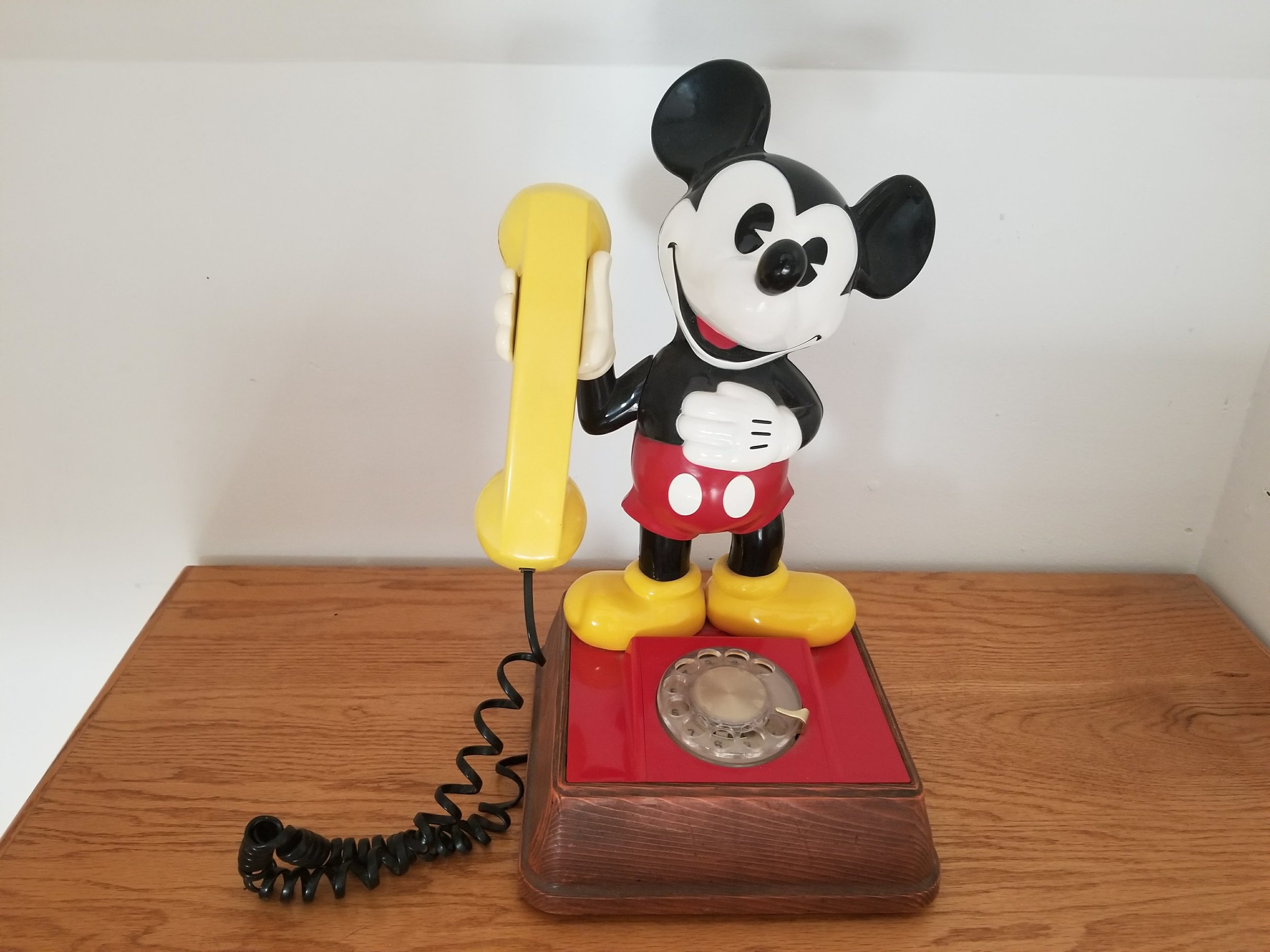 Mickey Mouse figure holding a yellow vintage rotary phone on a wooden surface.