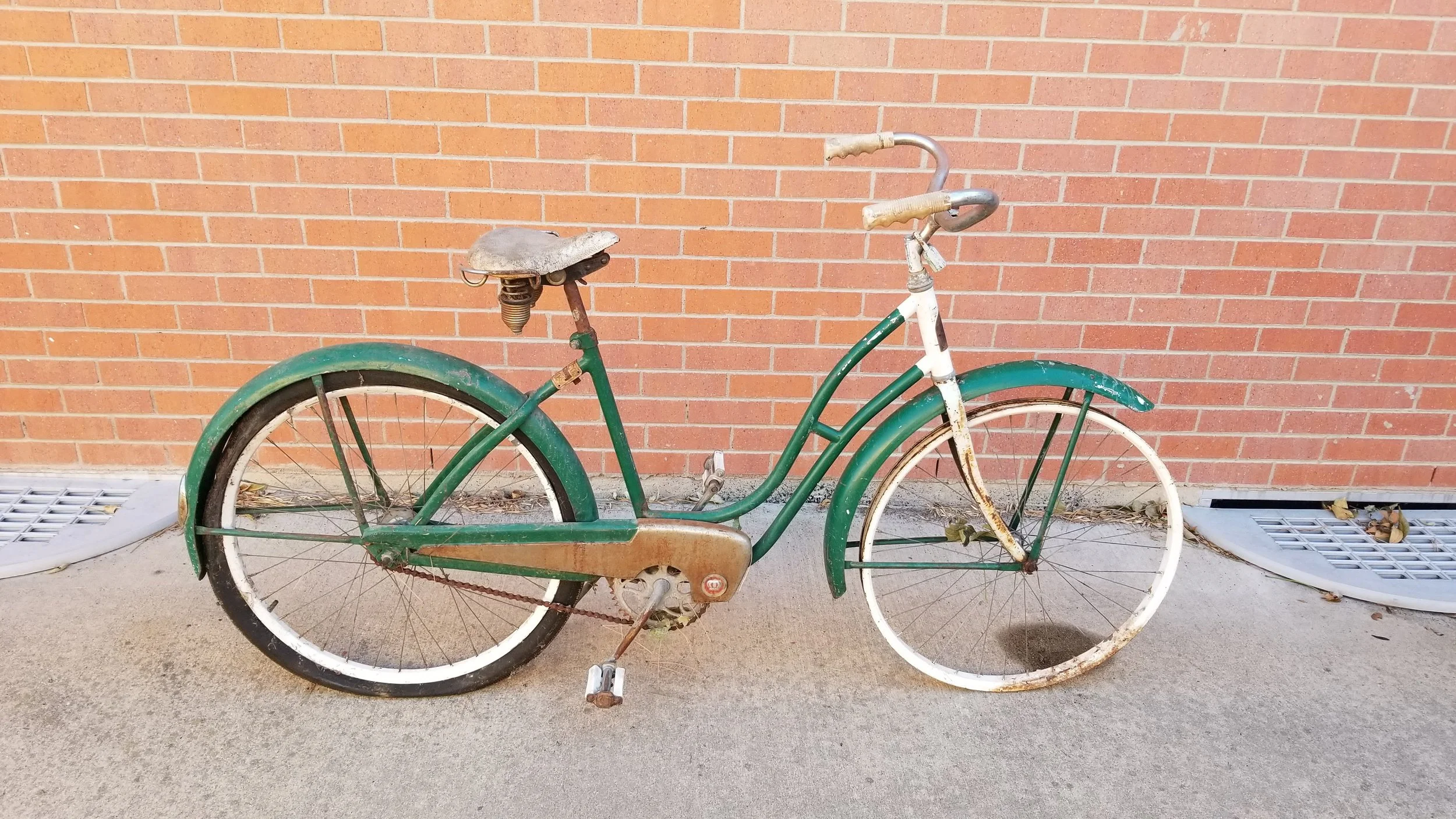 Old, rusty green and white bicycle with a brown seat leaning against a brick wall on a concrete ground.