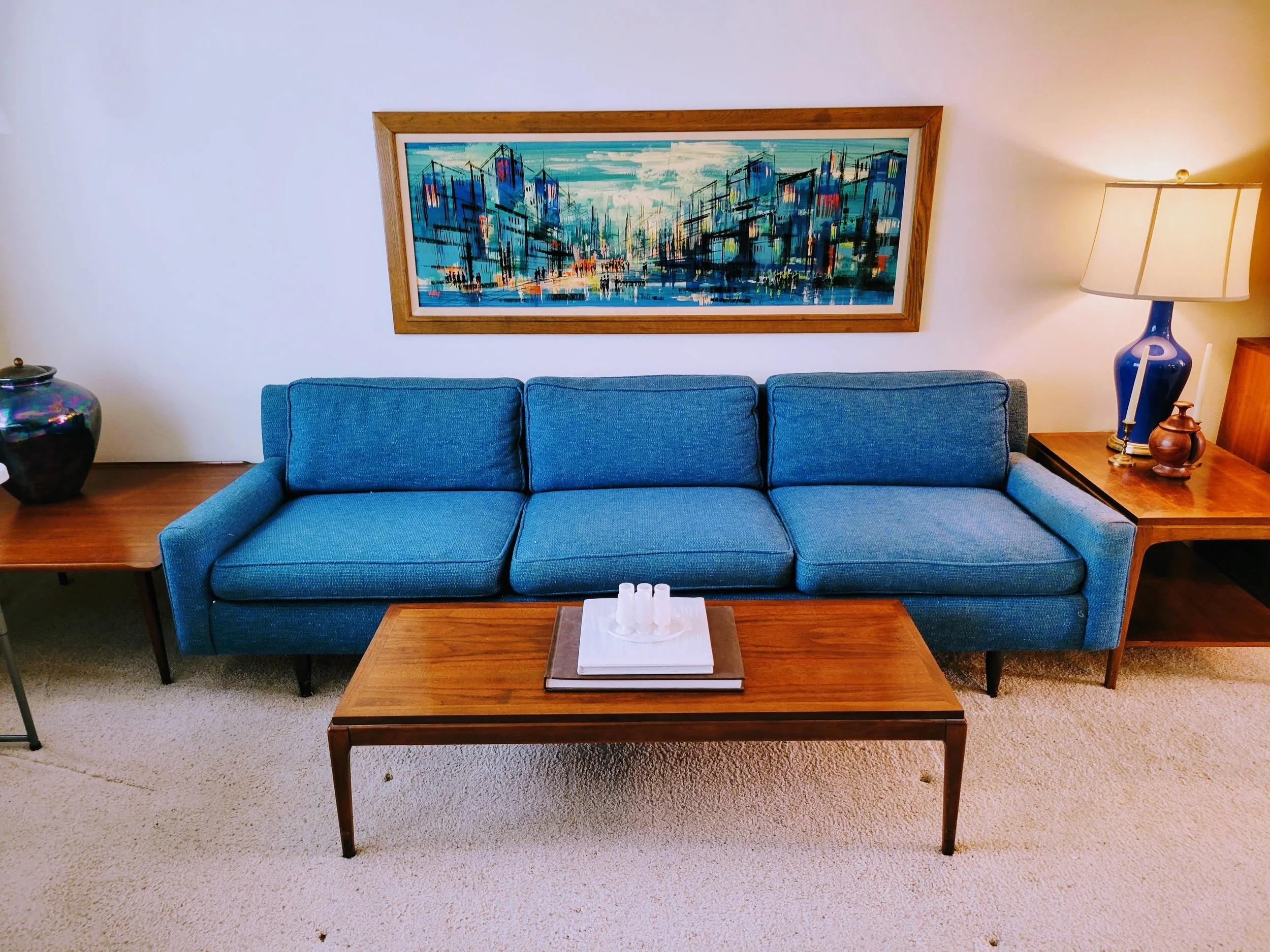 Living room with a blue sofa, wooden coffee table with white candles, abstract cityscape painting on the wall, table lamp with blue vase, and other decorative items.
