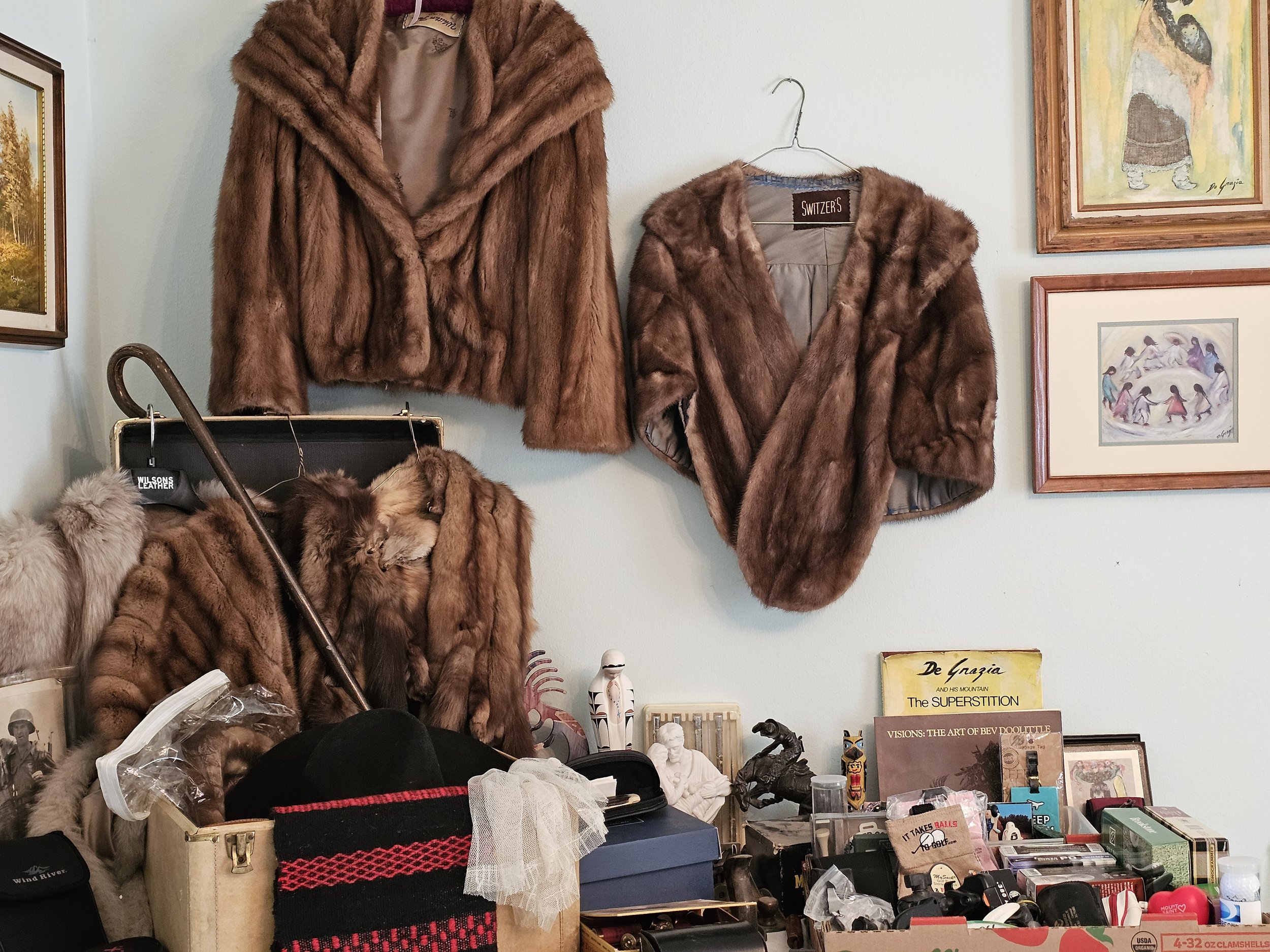Display of vintage fur coats, including a large brown fur coat hanging on a wall, a smaller brown fur jacket on a hanger, and a fur stole draped over a chair, with framed artwork on the wall and a cluttered table below.