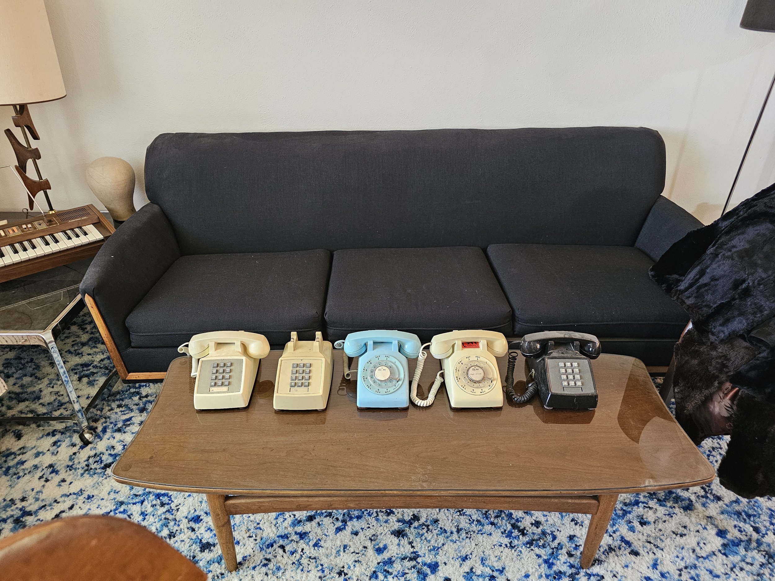 Five vintage rotary desk phones in various colors are lined up on a wooden coffee table in front of a black sofa in a living room.