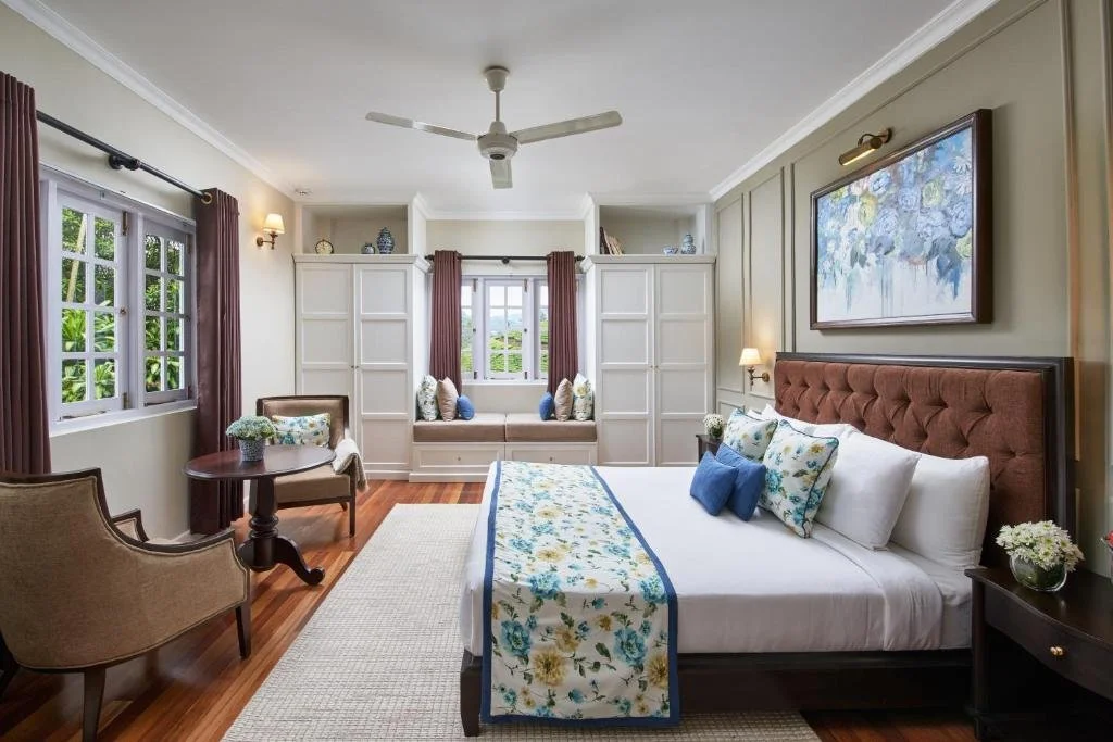 A cozy bedroom with a large bed, decorated with white and blue floral bedding and pillows. There are two nightstands with lamps and flowers, a window seat with cushions, a window with brown curtains, and a sitting area with two chairs and a small round table. The room has hardwood floors and beige walls.