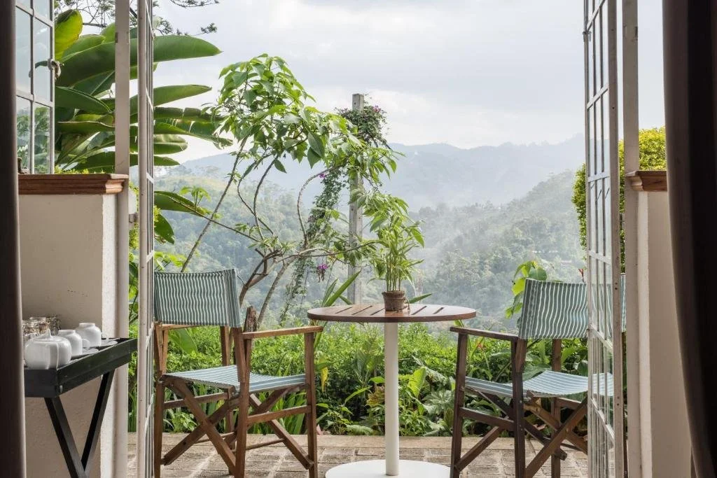 A cozy outdoor balcony with a small round table and two chairs, overlooking lush green mountains and clouds, with potted plants inside and tropical foliage outside.
