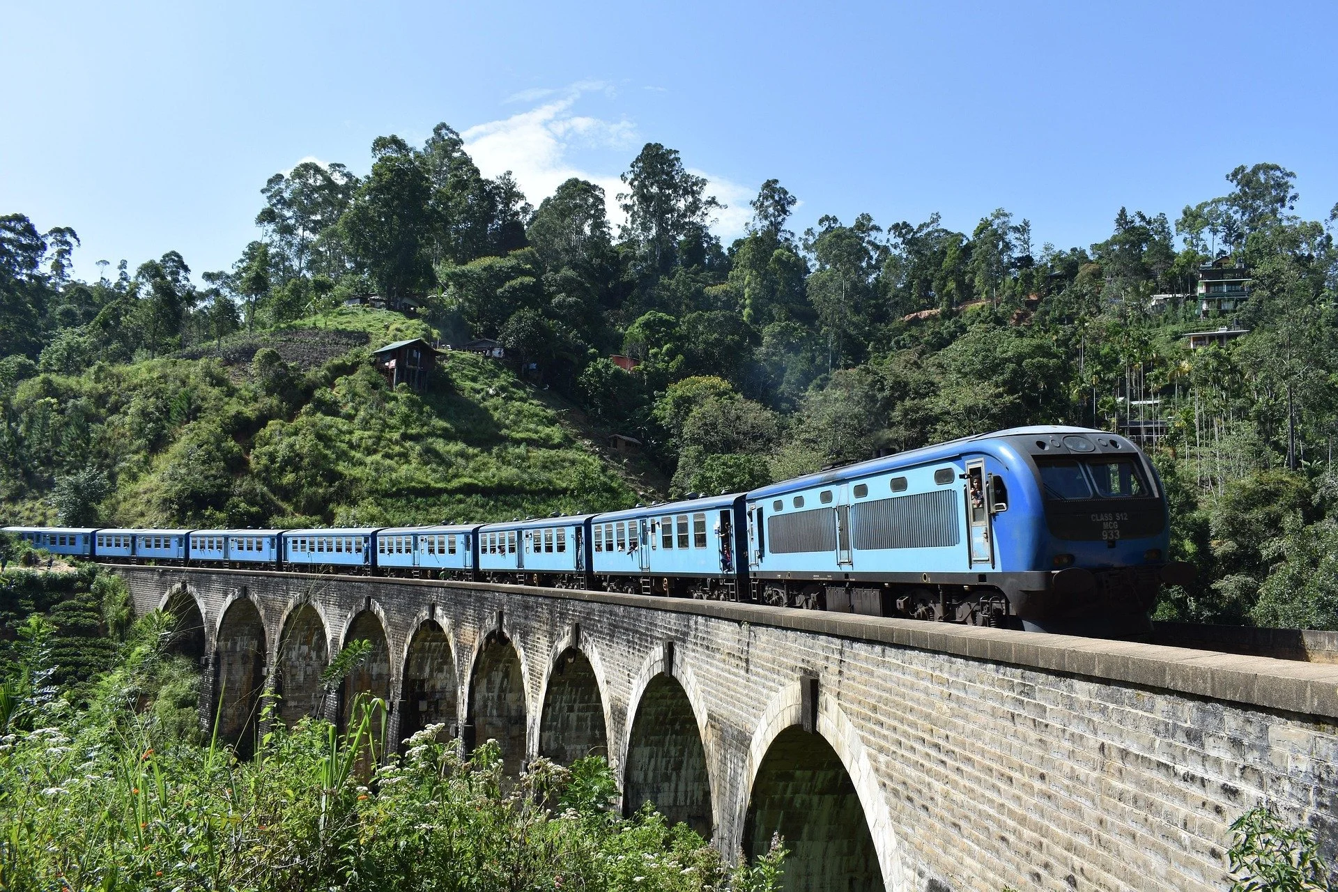 A blue passenger train traveling over a stone arch bridge in a green, hilly landscape with trees and houses