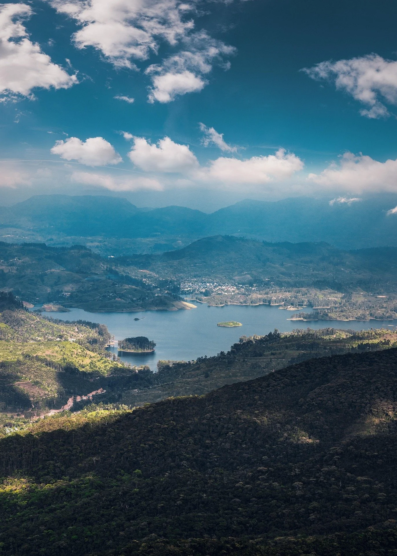 A scenic view of a large lake surrounded by green hills and mountains under a partly cloudy sky.