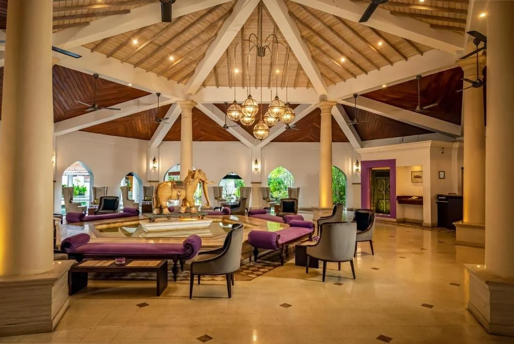 Elegant hotel lobby with a central round seating area around a water feature with a golden elephant sculpture. The space features high vaulted ceilings with wooden accents, chandeliers, and large windows with greenery outside.