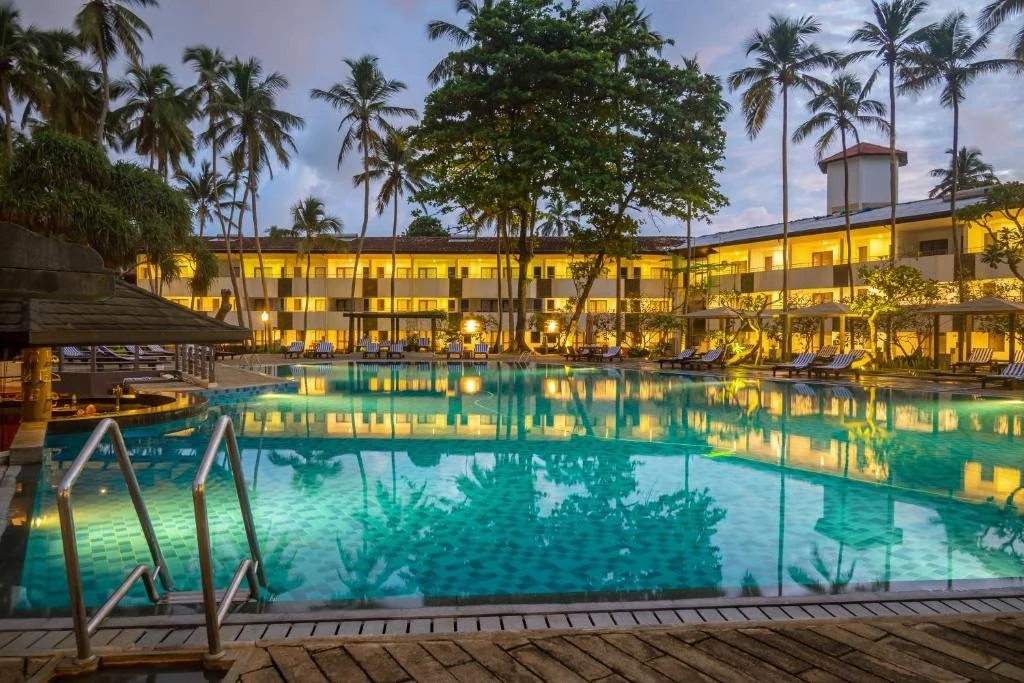 A pool area at a tropical resort with palm trees, lounge chairs, and a two-story building with yellow lights in the background during early evening.
