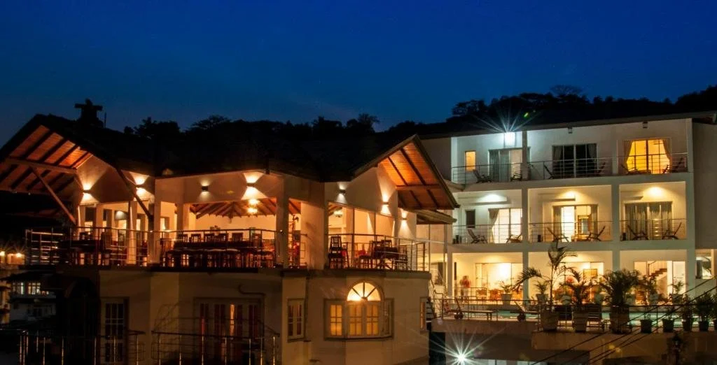 A multi-story hotel or resort building at night, with warm interior lights illuminating balconies and windows, surrounded by a dark sky and some trees in the background.