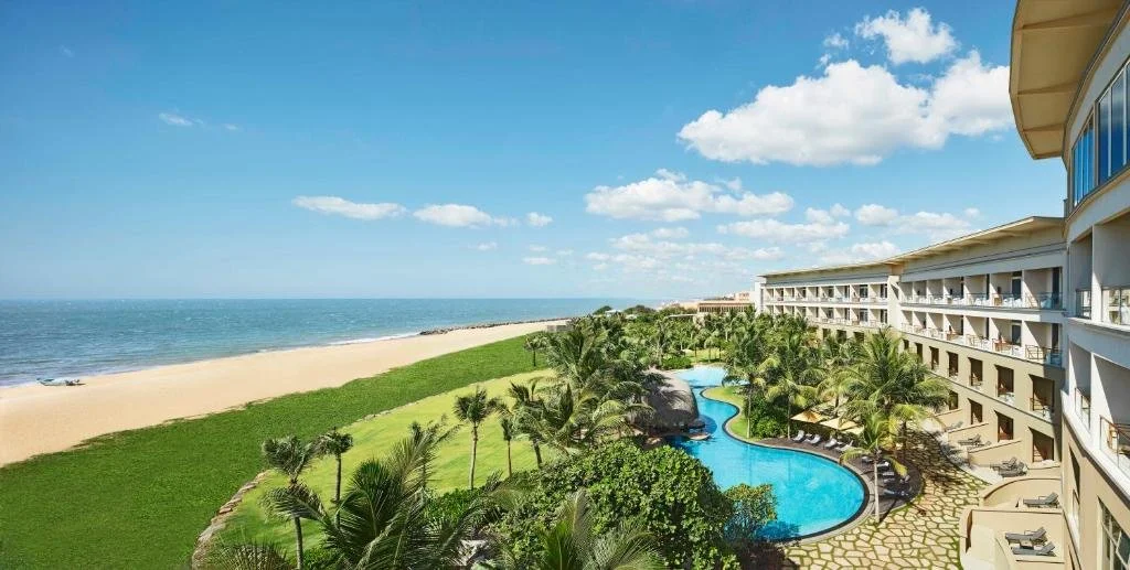 View of a beachfront resort with a swimming pool surrounded by palm trees, overlooking the beach and ocean under a blue sky with some clouds.