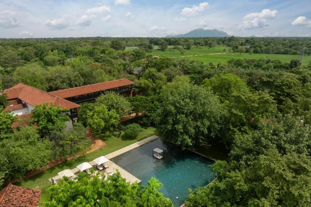 Aerial view of a lush green landscape with trees, a swimming pool, buildings with red-tiled roofs, and distant mountains under a partly cloudy sky.
