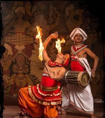 Two performers in traditional African attire perform a fire dance, one holding fire on their hand while the other beats a drum. They are in front of a patterned background.