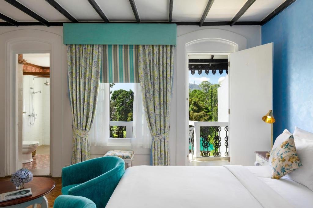 A bright hotel room with white walls, blue accent wall, and a balcony door overlooking greenery. The room has a bed with white bedding and floral pillows, a turquoise armchair, and colorful patterned curtains.