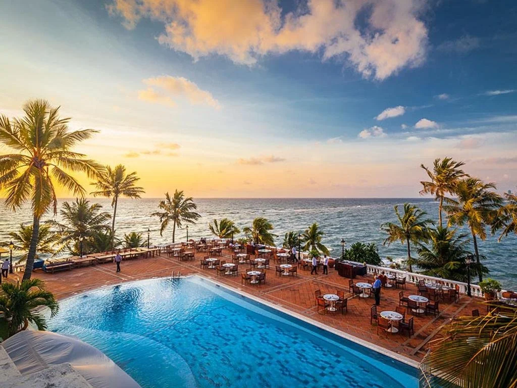 Poolside view with ocean and sunset, surrounded by palm trees and outdoor dining tables.