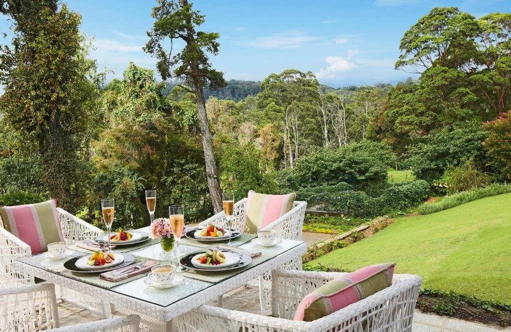 An outdoor dining table set up on a patio with six plates of food, six champagne glasses, and a small flower arrangement, overlooking a lush green landscape with trees and a grassy slope under a partly cloudy sky.