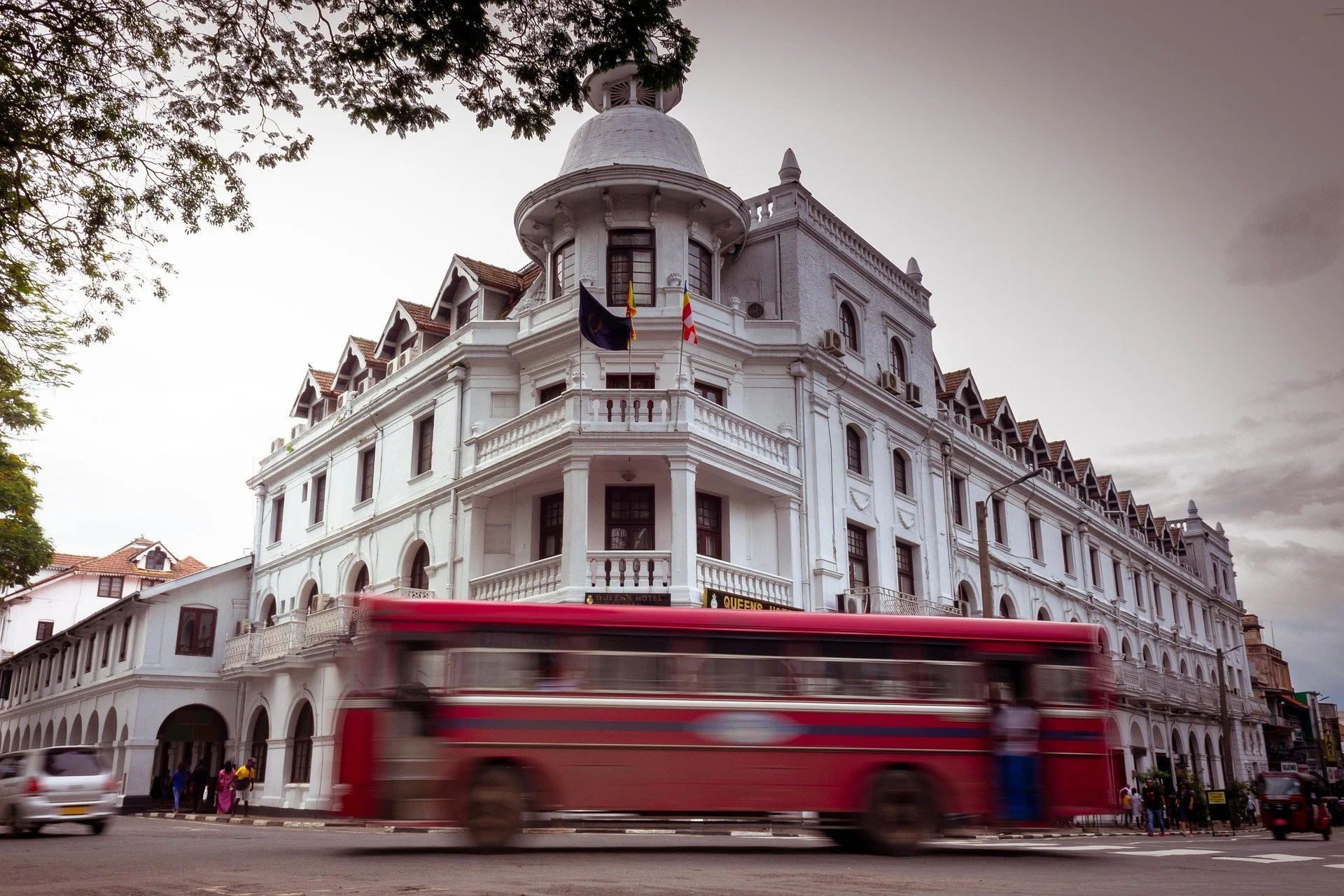 A white colonial-style building with multiple windows, balconies, and a domed tower, with a red bus passing in front and a few pedestrians on the street.