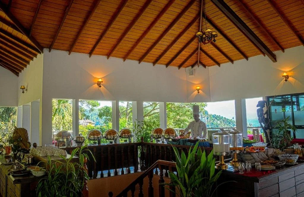 Buffet setup with chafing dishes, glasses, and food items in a room with large windows, wooden ceiling, and a chef preparing food.