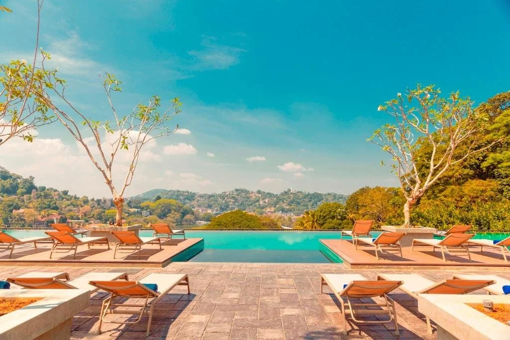 A luxurious outdoor swimming pool area with lounge chairs, surrounded by trees and a scenic view of distant hills under a bright blue sky.